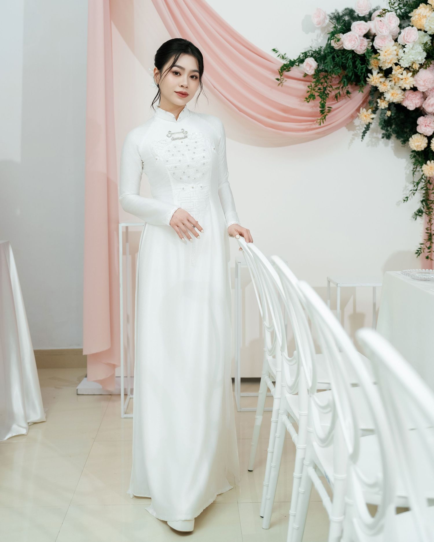 Traditional Vietnamese white ao dai dress with floral decor and pink drapery in an elegant wedding setting.