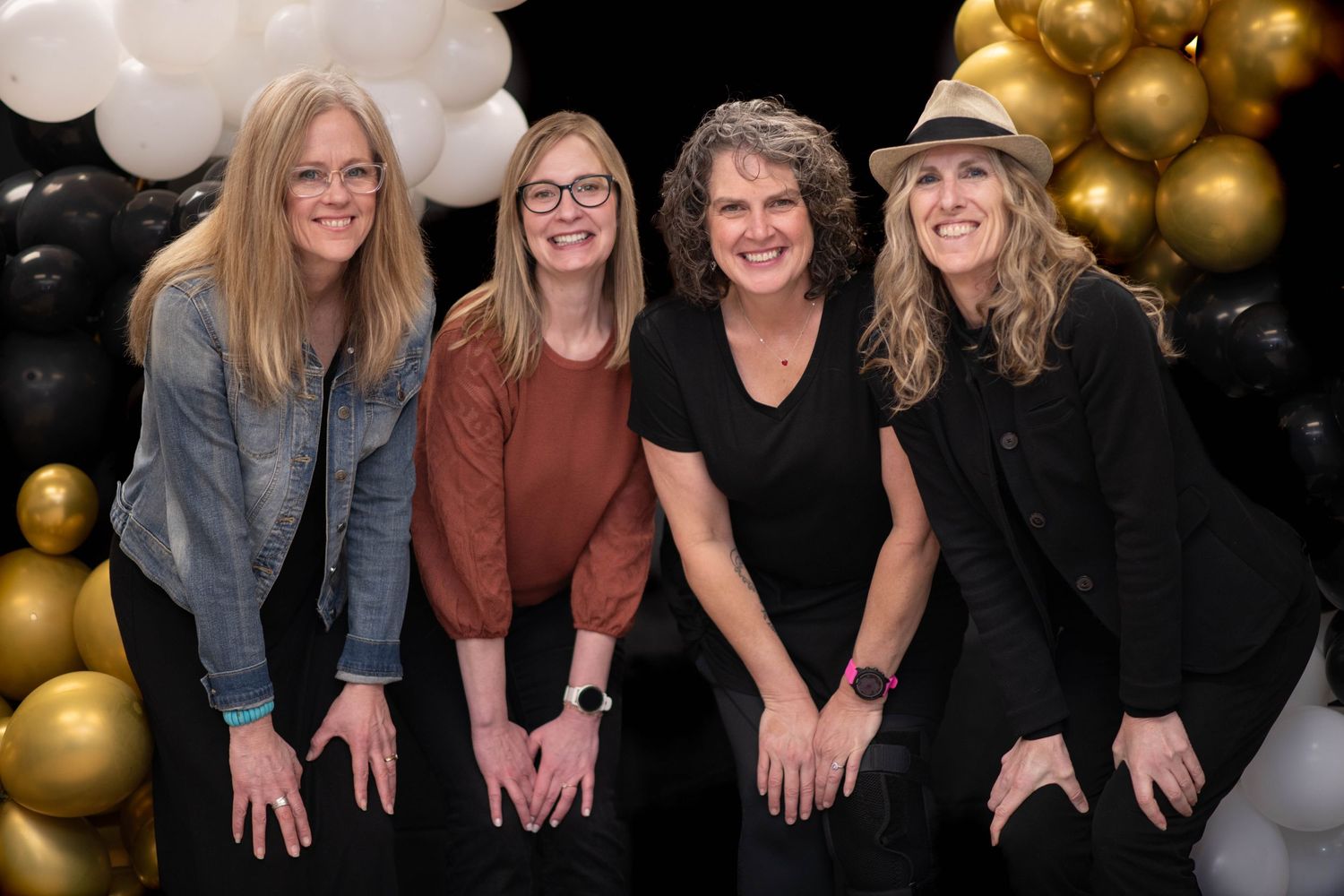 Four women smile together in front of black, gold, and white balloon backdrop.