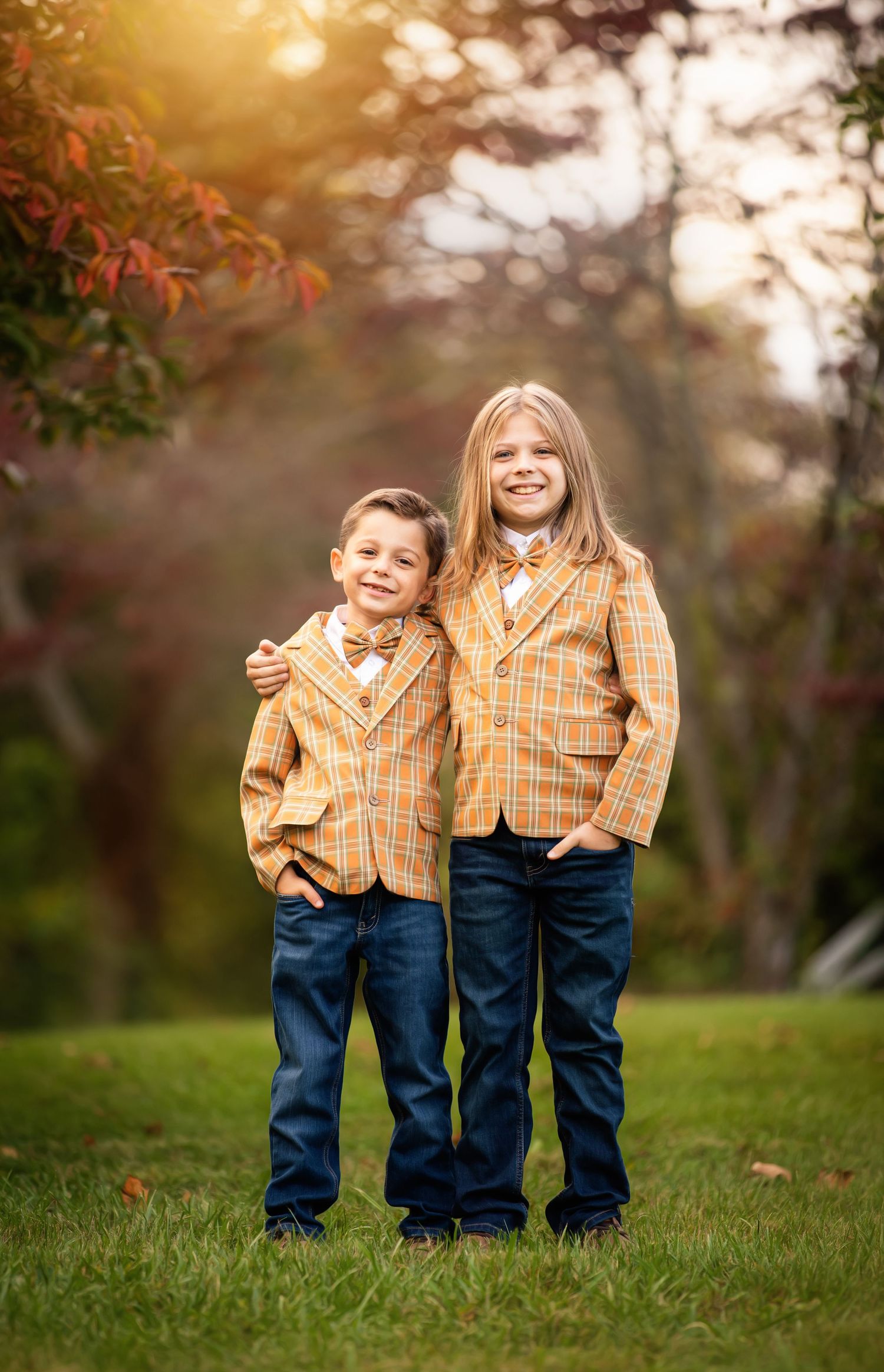 Two children wearing matching orange plaid blazers and jeans stand together on grass during golden hour sunset.