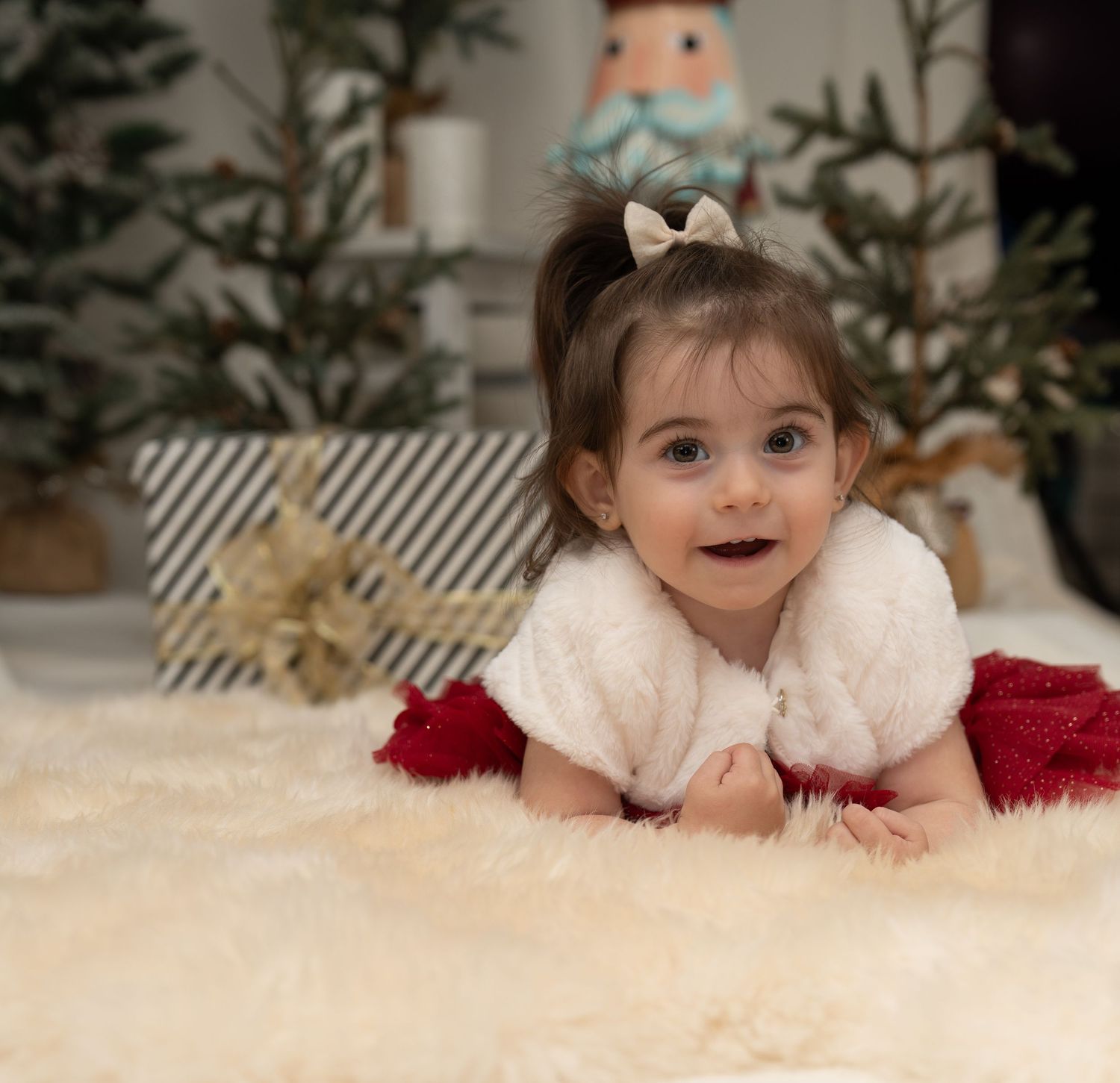 Baby in white dress lying on fluffy white carpet near Christmas decorations.