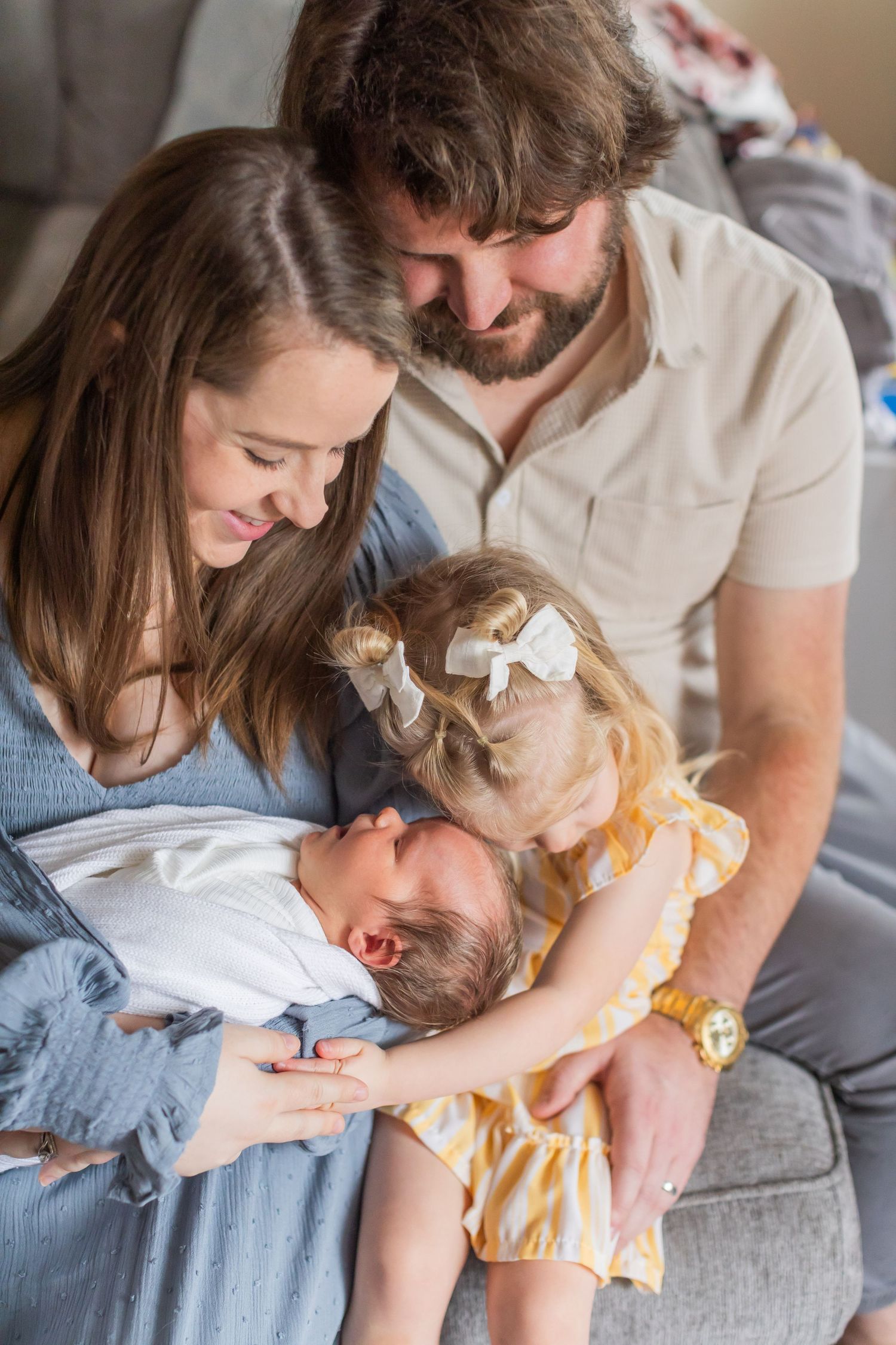 A family shares a tender moment together on a gray couch while admiring a newborn baby at home.