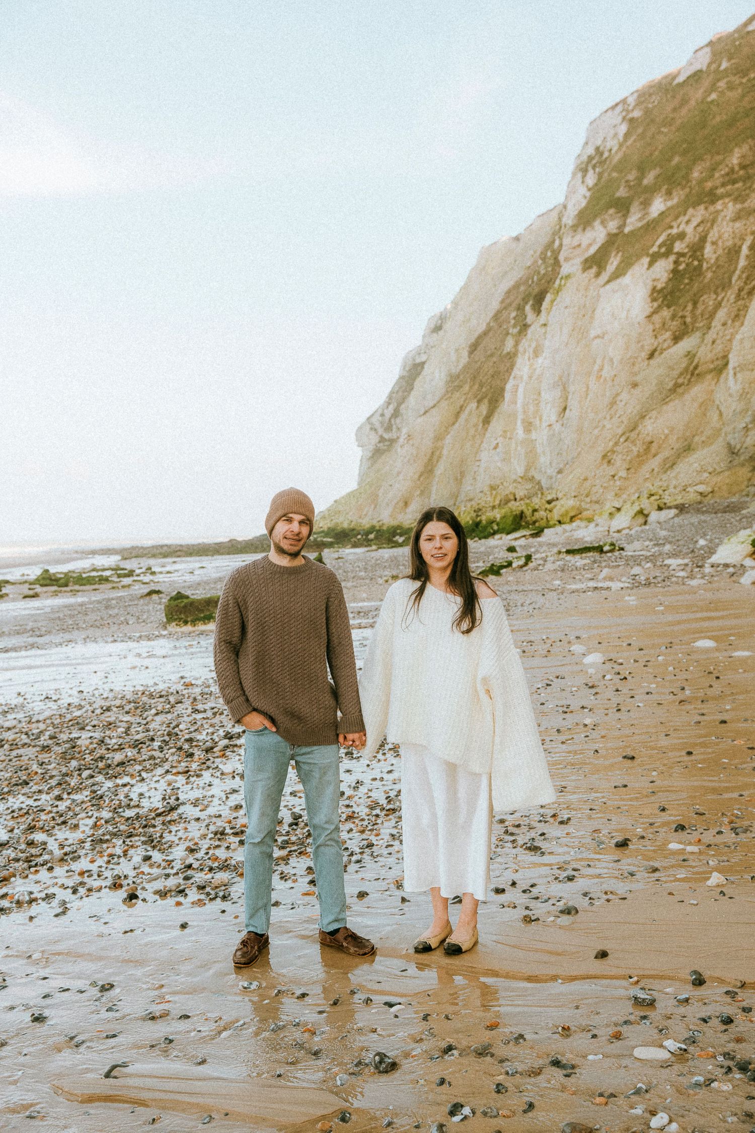 A couple stands together on a rocky beach with tall white cliffs in the background on an overcast day.