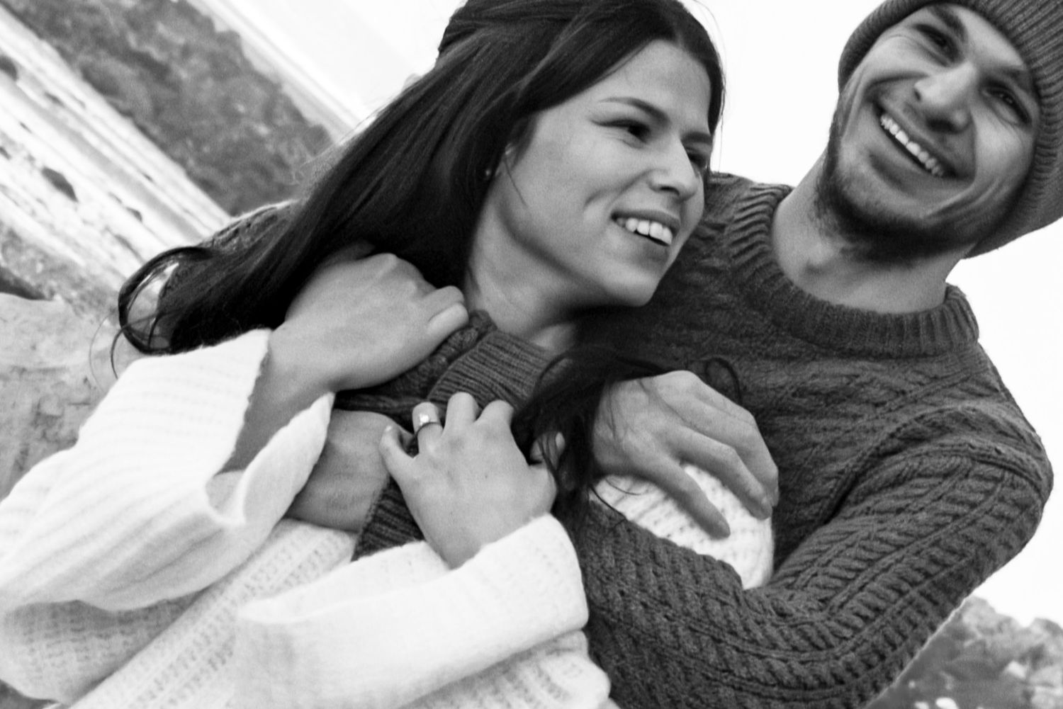 Black and white photo of a happy couple laughing and embracing outdoors while wearing winter sweaters.