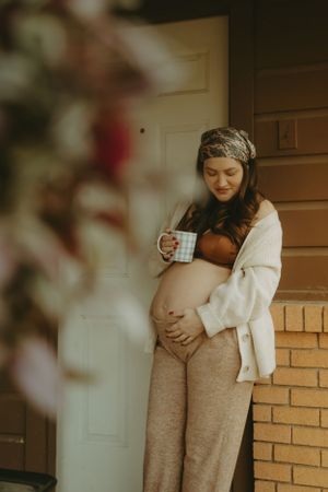 Expectant mother in cozy attire holds coffee mug while standing on front porch in warm lighting.