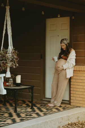 Pregnant woman relaxes on decorated porch with macrame plant hanger and geometric rug.
