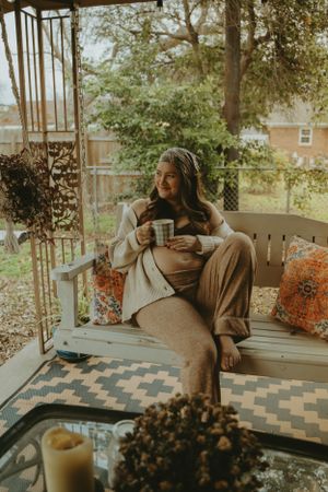 Cozy autumn maternity scene with woman relaxing on porch swing holding coffee.