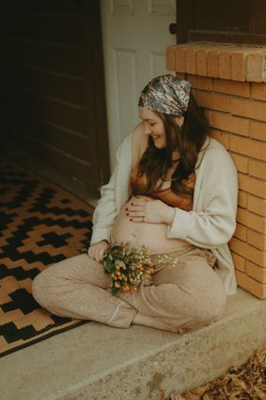 A maternity portrait of a woman in comfortable clothing holding dried flowers on a porch step.