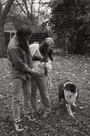 A black and white maternity photo with a couple and their dog in a leaf-covered yard.
