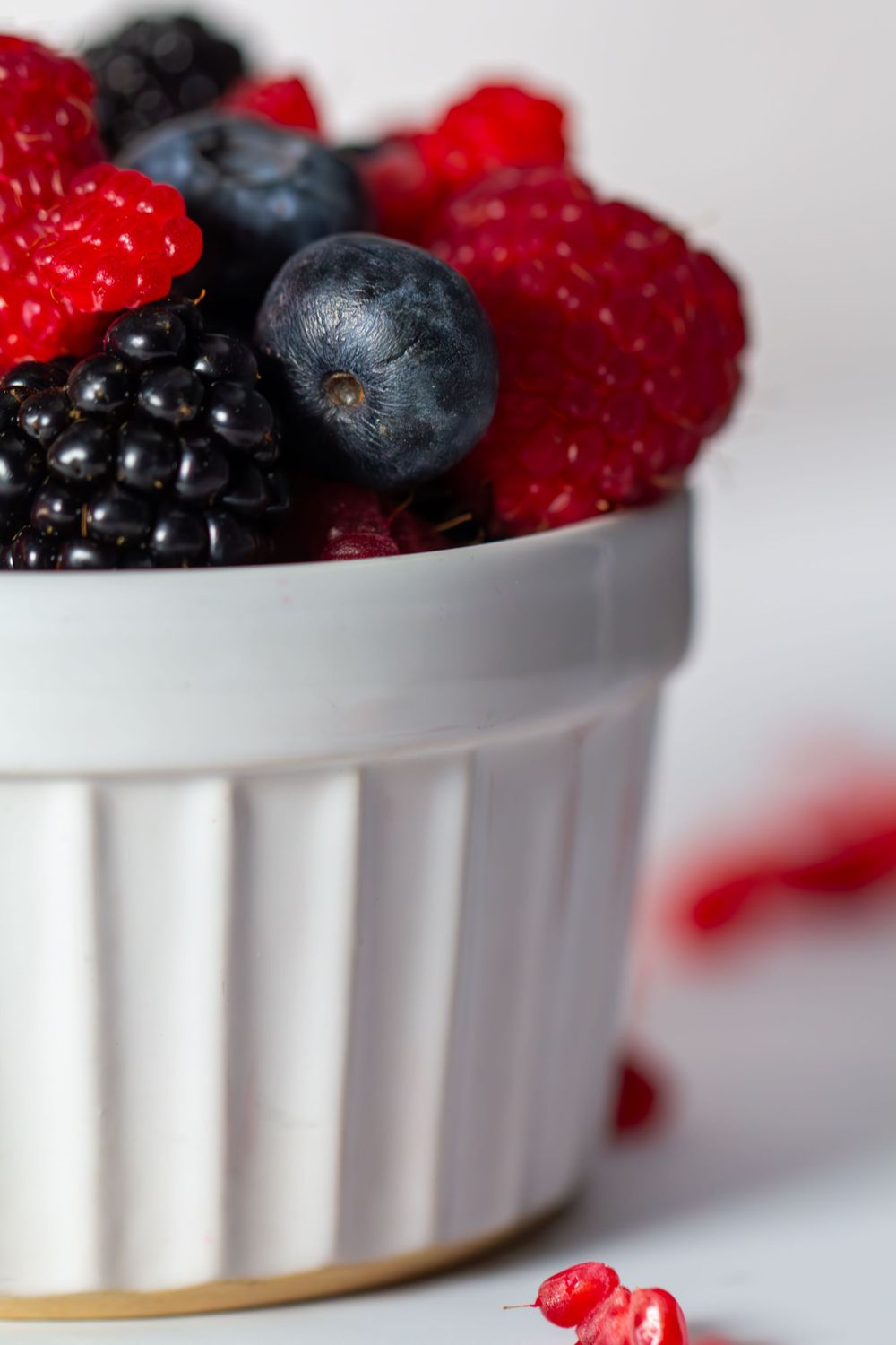 Fresh raspberries, blueberries and blackberries in a white ceramic ramekin against a white background with scattered raspberry crumbs.