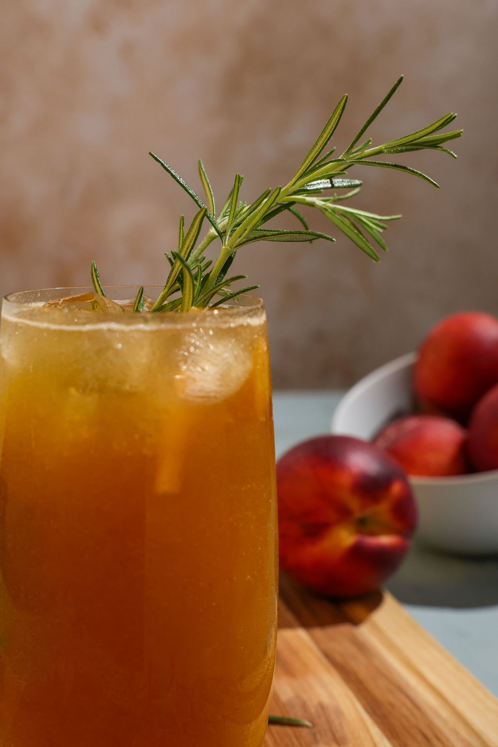 A refreshing peach iced beverage garnished with fresh rosemary sprig, with whole peaches visible in background.