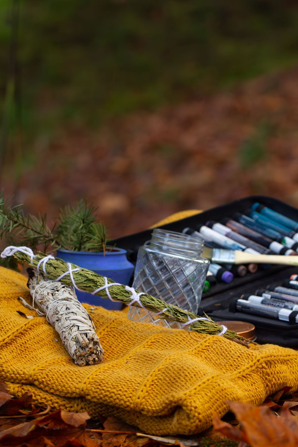 Sage smudge stick rests on yellow knit fabric alongside Native American Artist's art supplies and crystal glass in autumn leaves.
