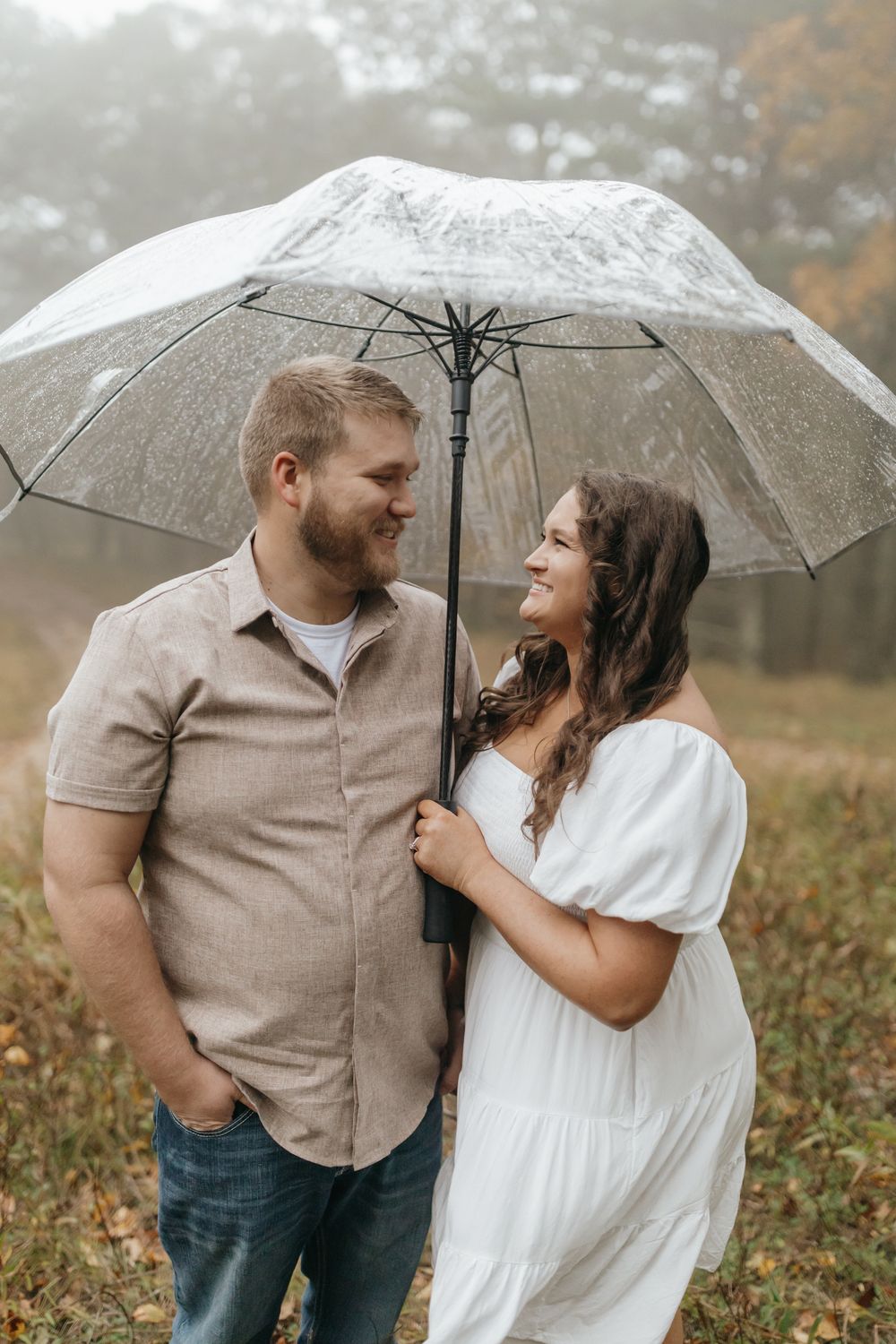 A couple shares a tender moment under a clear umbrella on a misty autumn day.