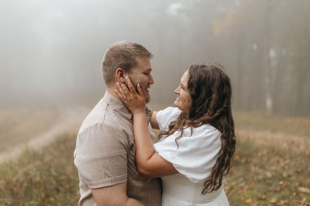 A tender moment between a couple in white and beige outfits against a foggy outdoor backdrop.