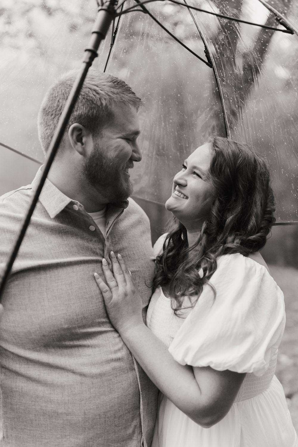 A black and white portrait of a couple sharing an intimate moment under a clear umbrella.