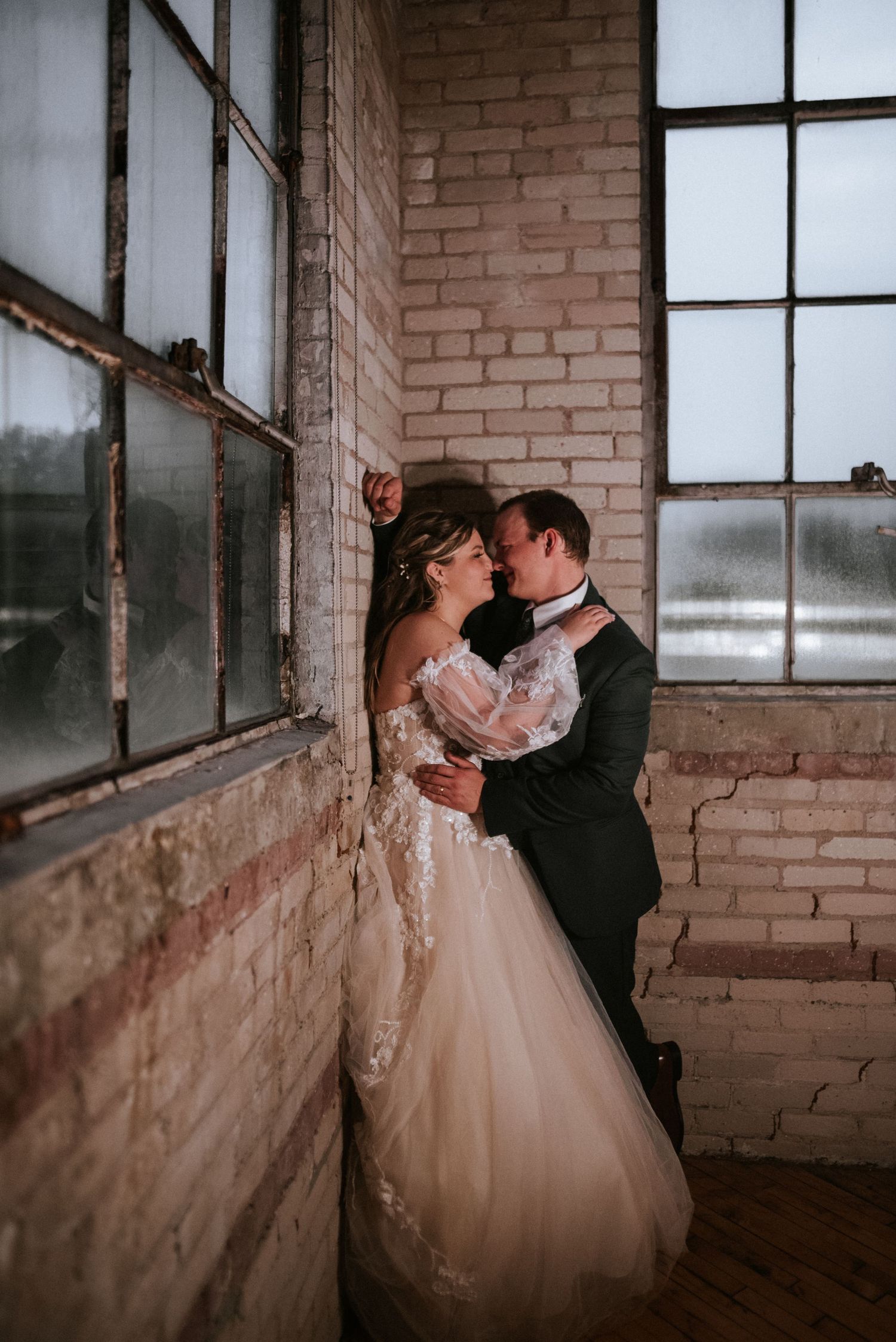 A romantic wedding moment captured in an industrial brick building with large windows and soft natural light.