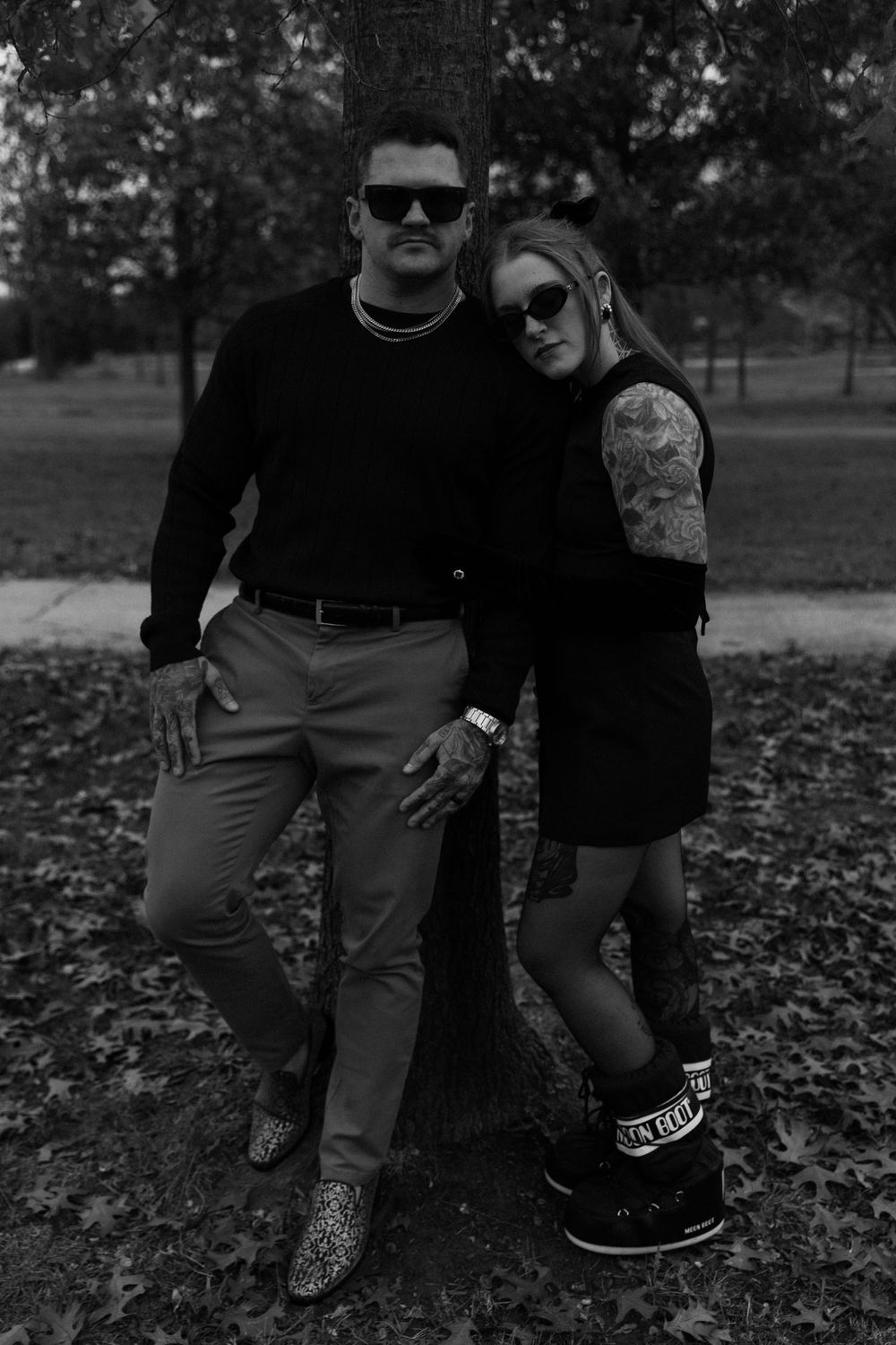 A fashionable couple in black attire poses against a tree in a park during autumn.