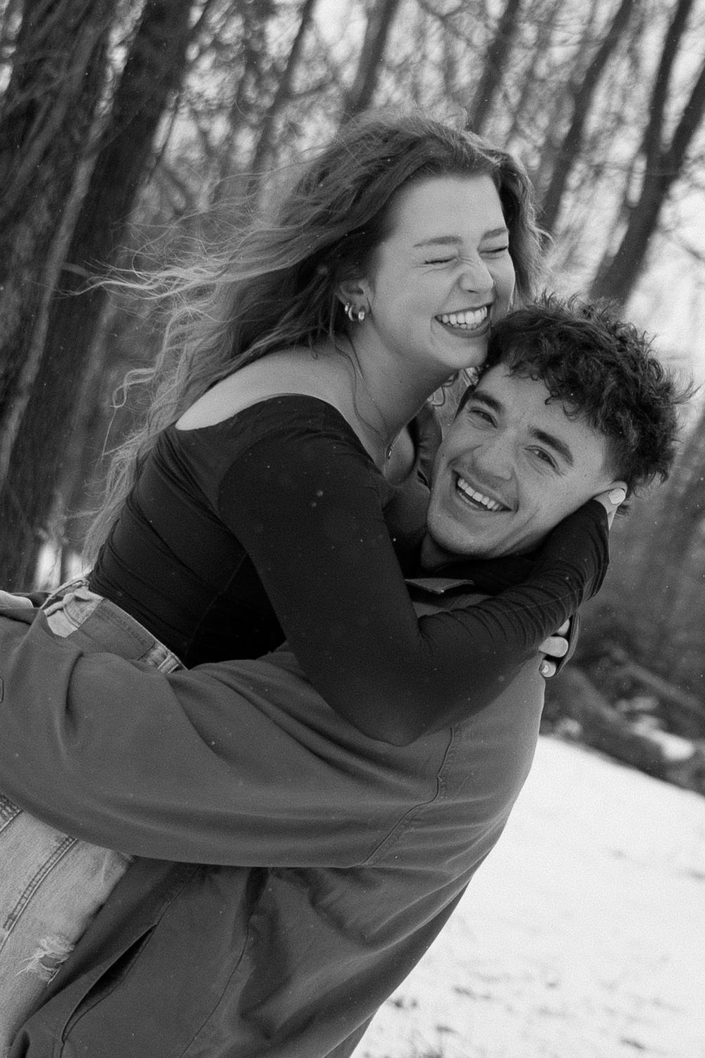 A joyful couple sharing laughter and embrace in a snowy forest setting captured in black and white.