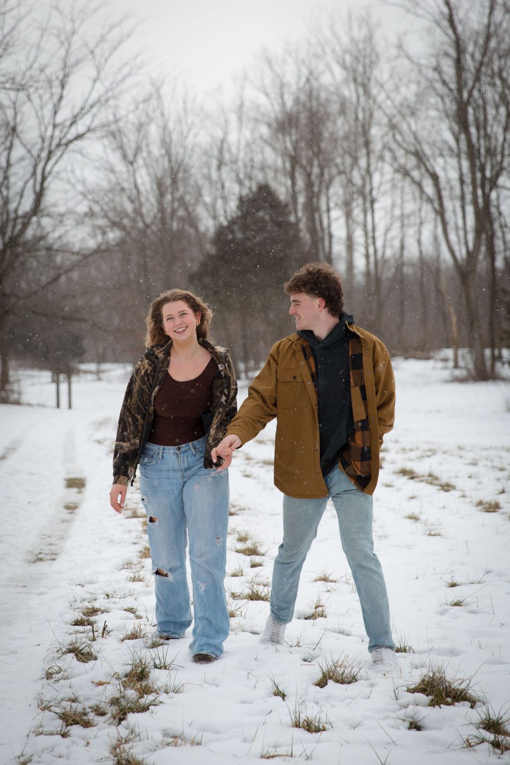 Couple holding hands while walking through snow-covered landscape with bare trees.
