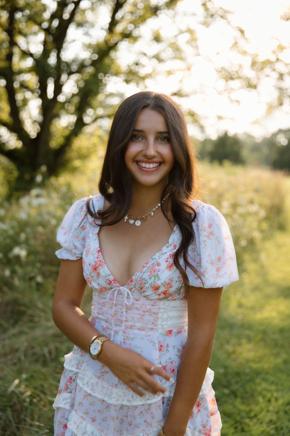 A woman in a floral summer dress stands outdoors in soft golden sunlight surrounded by greenery.