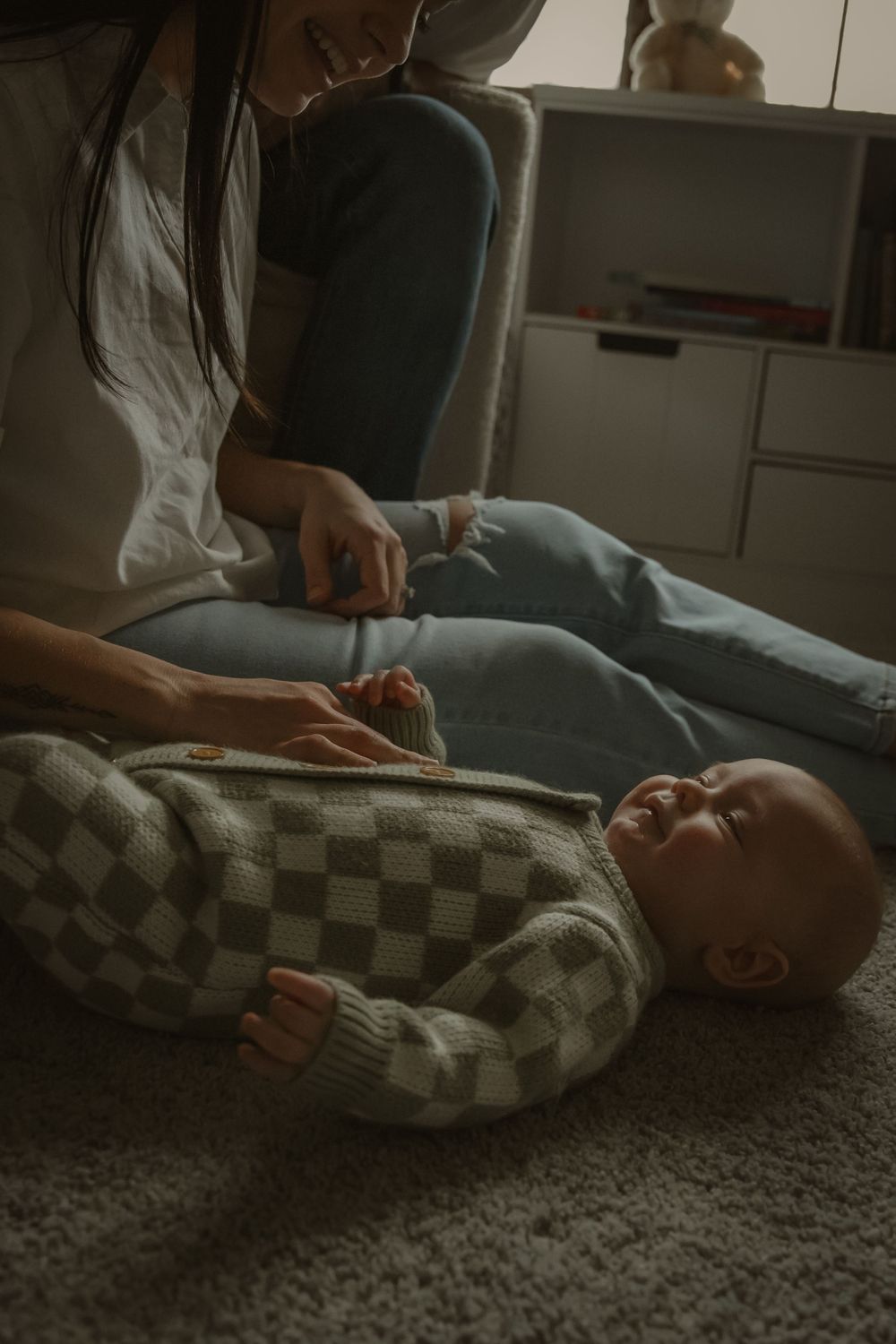 A baby in a checkered sweater lies on a carpet while receiving gentle care in warm, dim lighting.