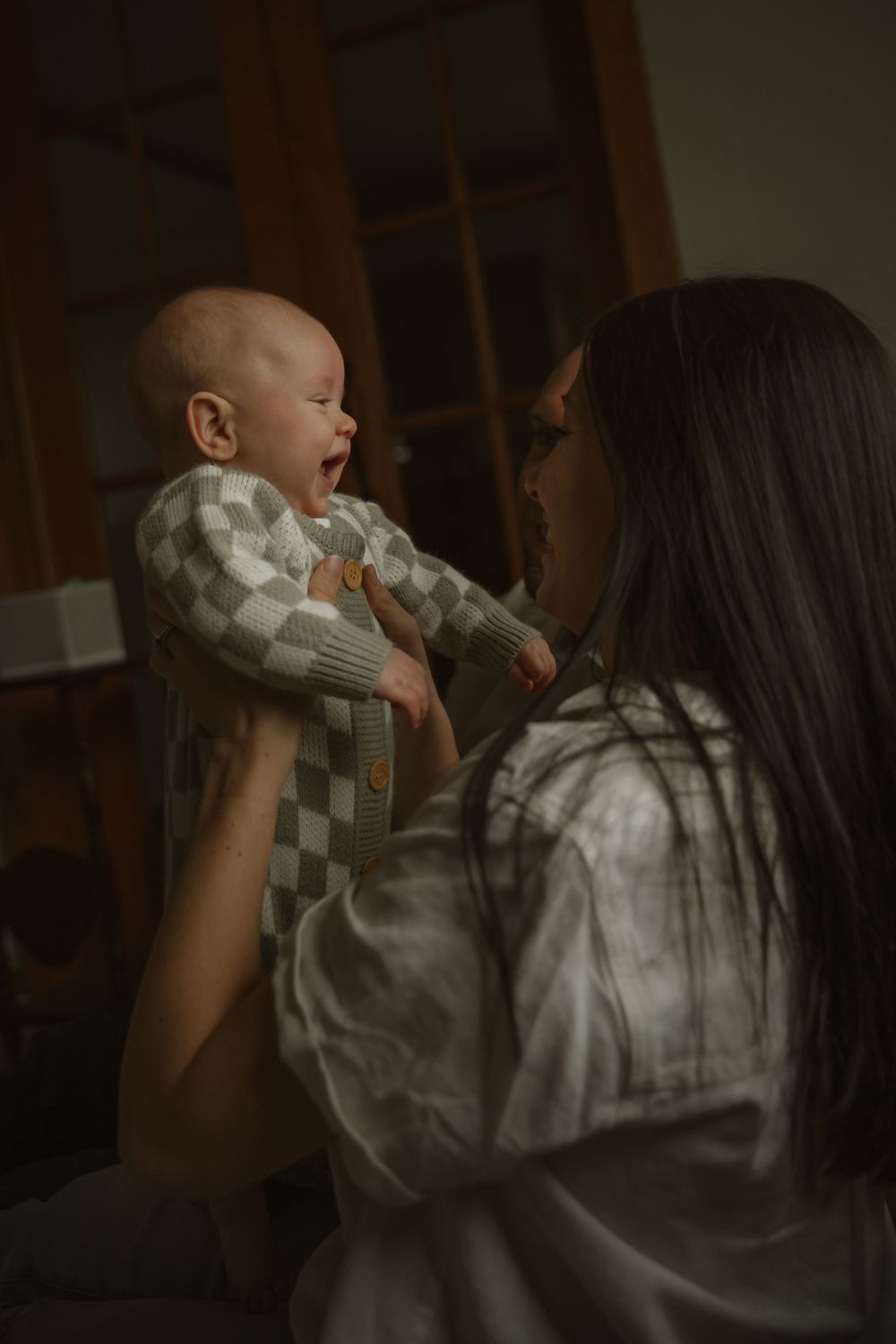 Baby in checkered outfit smiles while being held up in dim lighting with warm moody atmosphere.