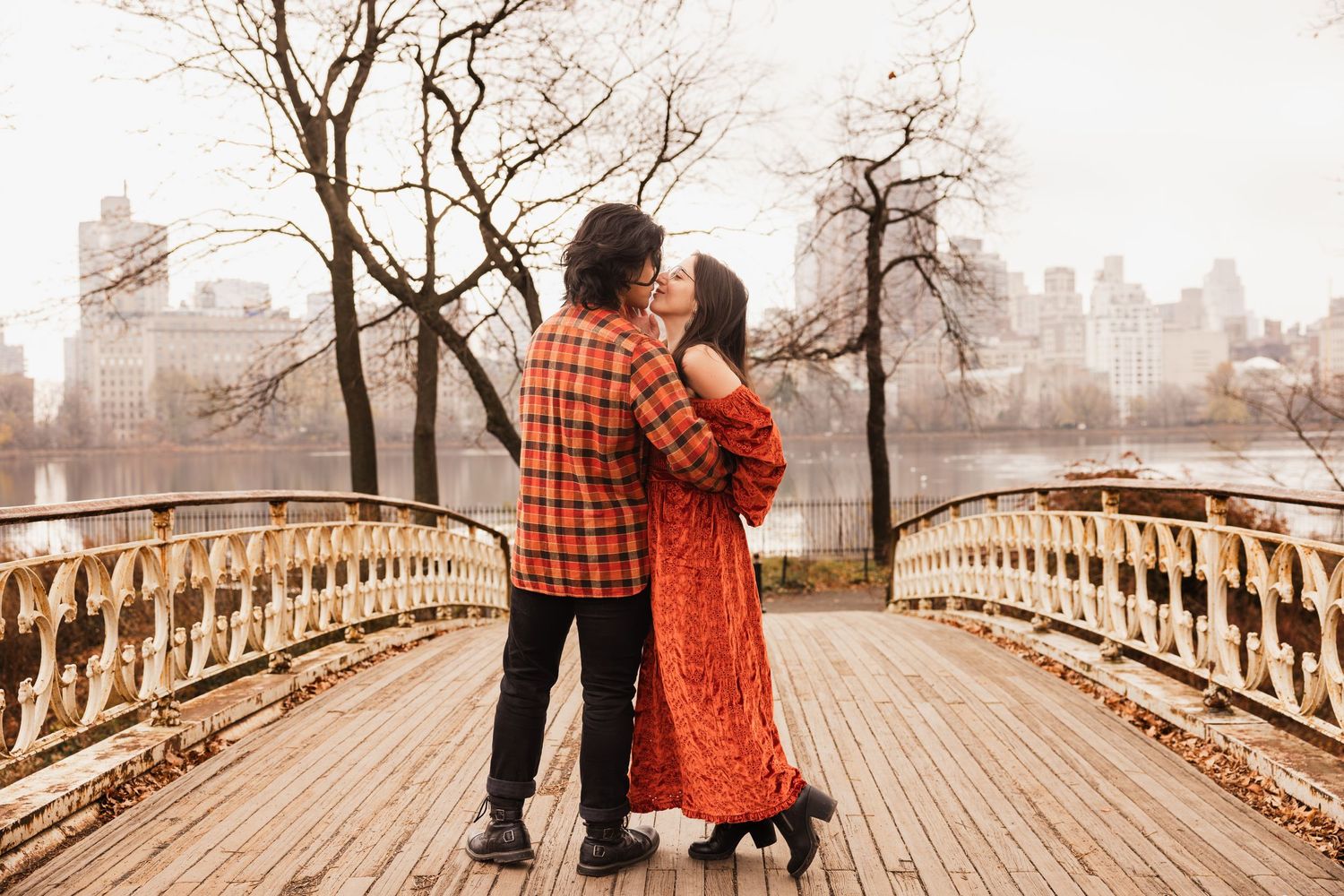 A couple embraces on a bridge in Central Park with NYC skyline in background during autumn.