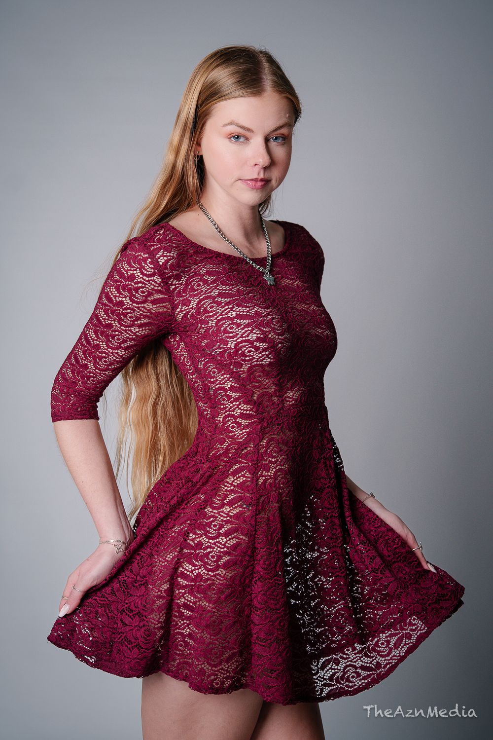 Model wearing burgundy lace dress twirls in studio setting against grey background.