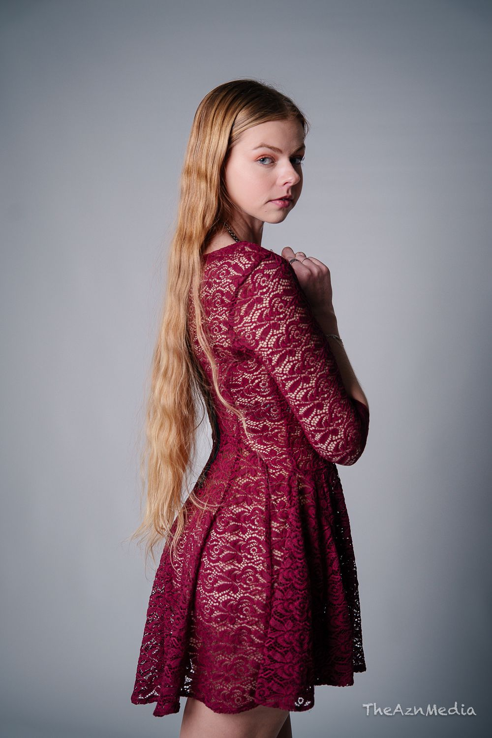 Long-haired model poses in burgundy lace dress against grey backdrop during fashion photoshoot.