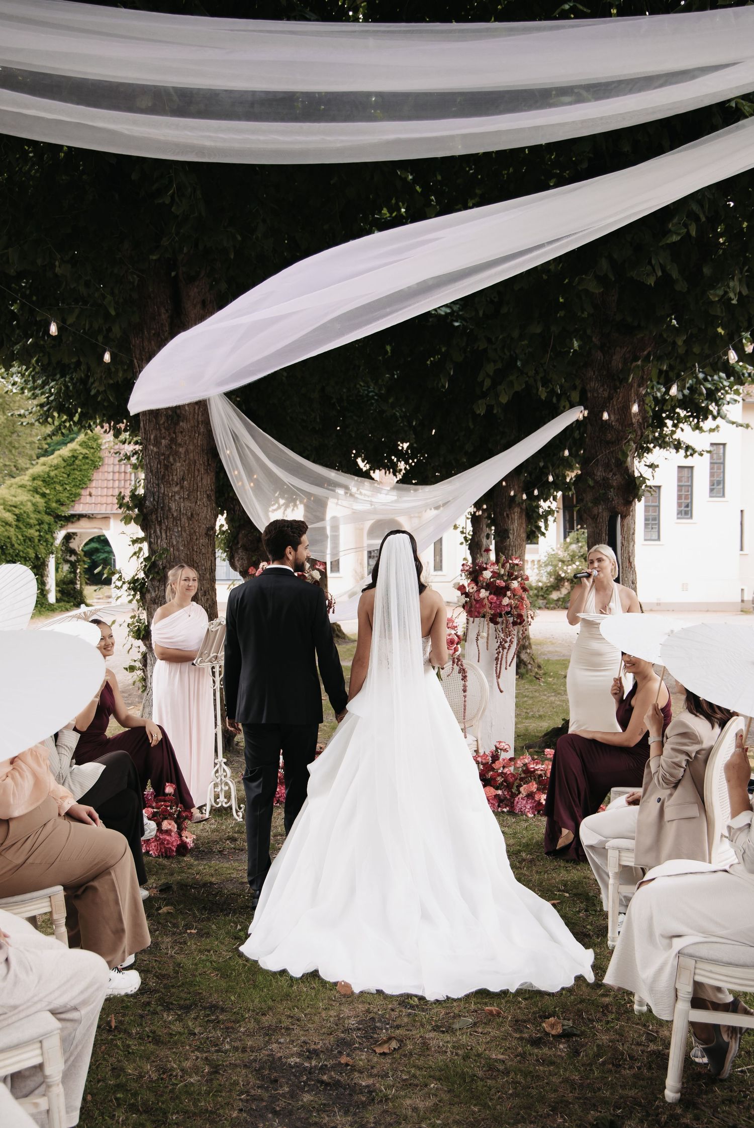 Bride and groom walk down outdoor aisle under flowing white fabric canopy, guests seated beside floral arrangements.