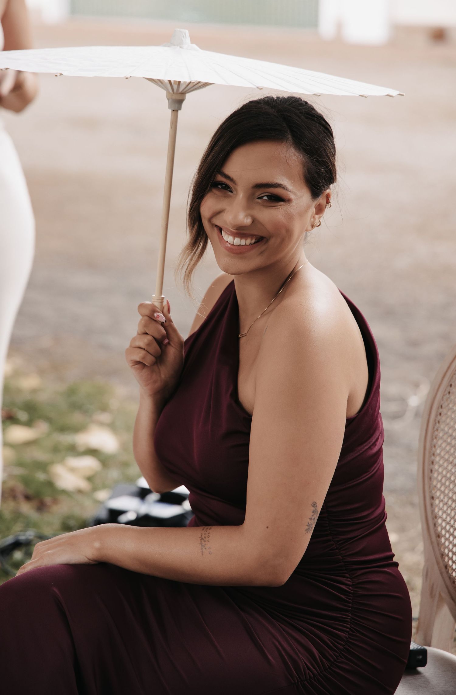 Smiling woman in burgundy one-shoulder dress holding a white parasol umbrella at an outdoor event.