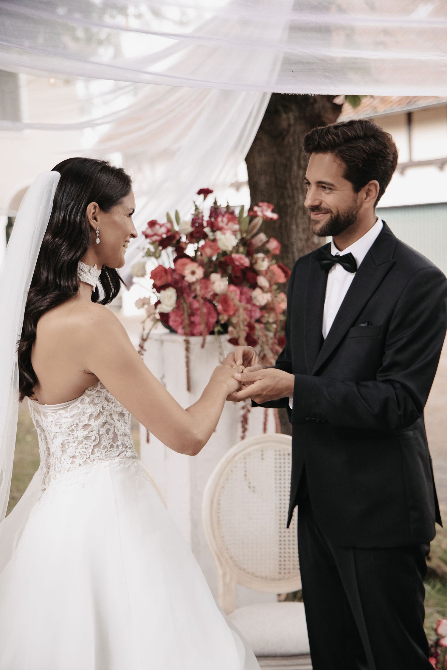 Bride and groom exchanging rings at outdoor wedding ceremony, surrounded by floral arrangements.