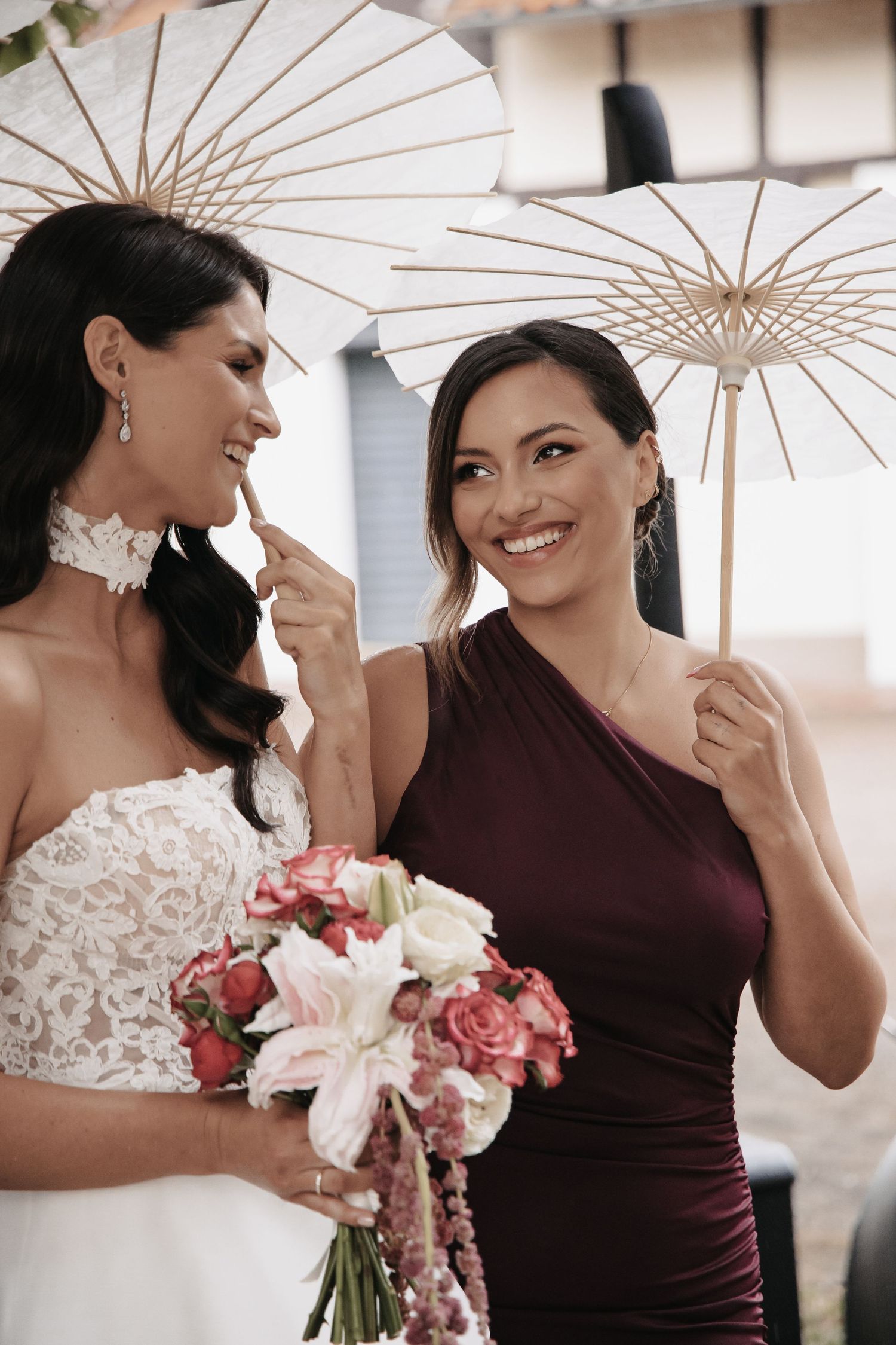 Bride in white lace gown and bridesmaid in burgundy dress holding parasols and a floral bouquet, smiling together.