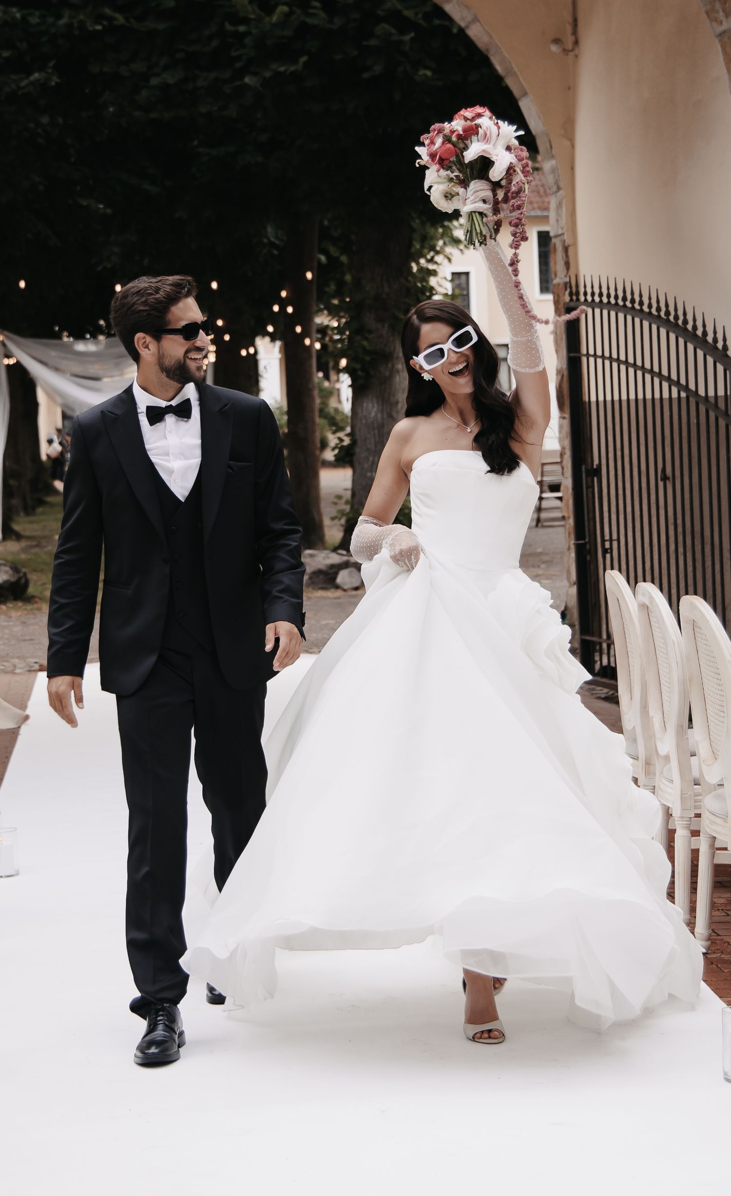 Joyful bride in white gown and sunglasses raises bouquet while walking with groom in black tuxedo at outdoor wedding.