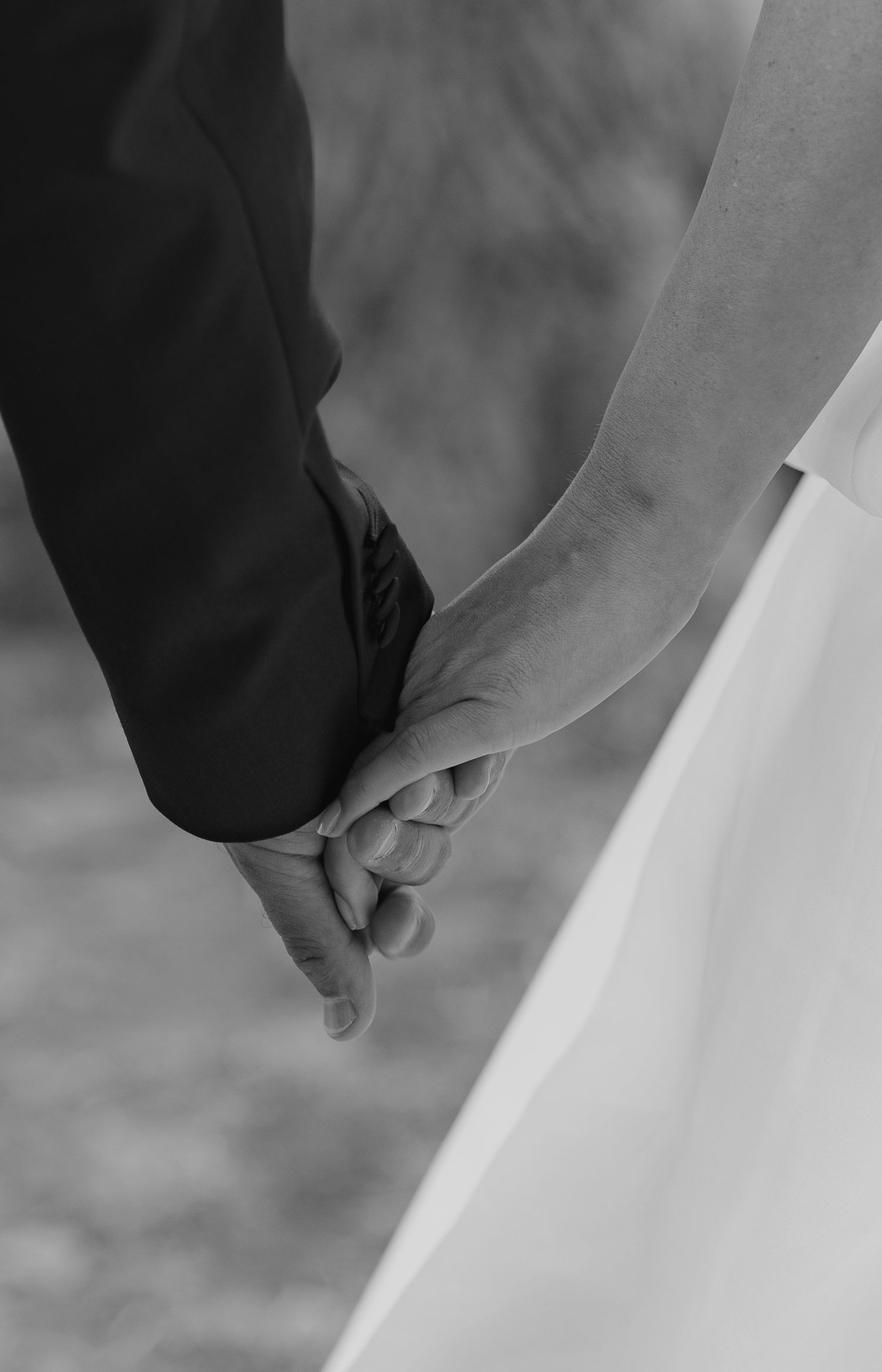 Black and white photo of a bride and groom holding hands, showcasing wedding rings and elegant attire.