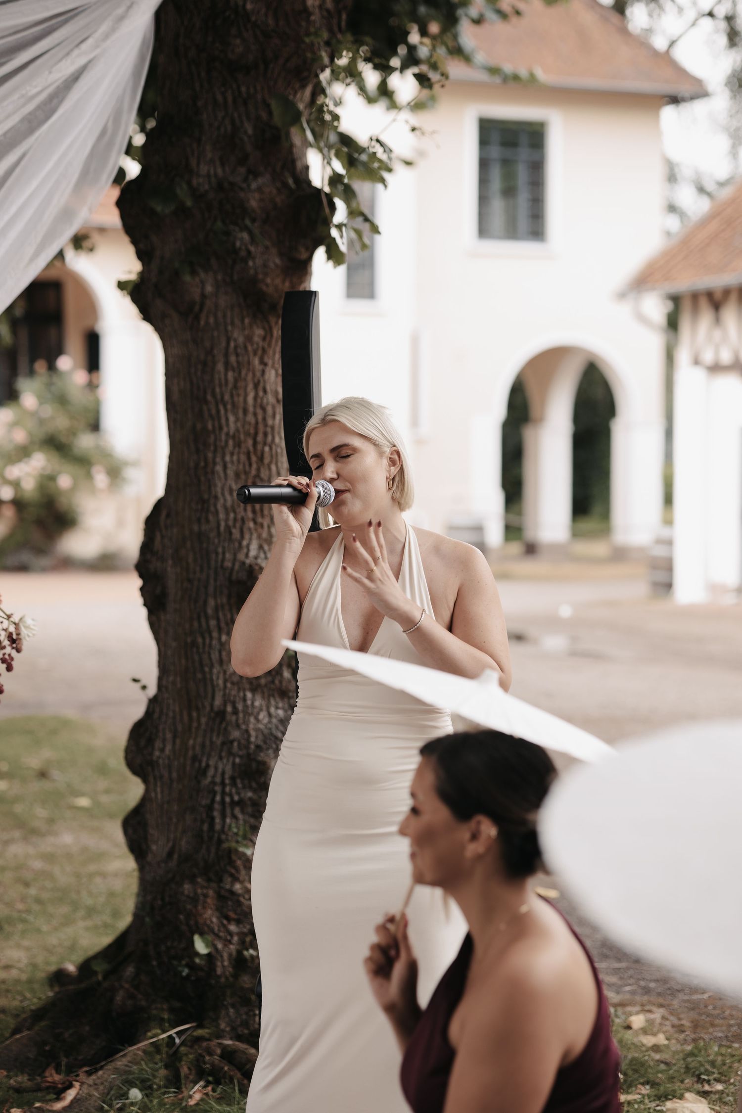 Blonde bridesmaid in white halter dress singing into microphone outdoors at elegant wedding venue.