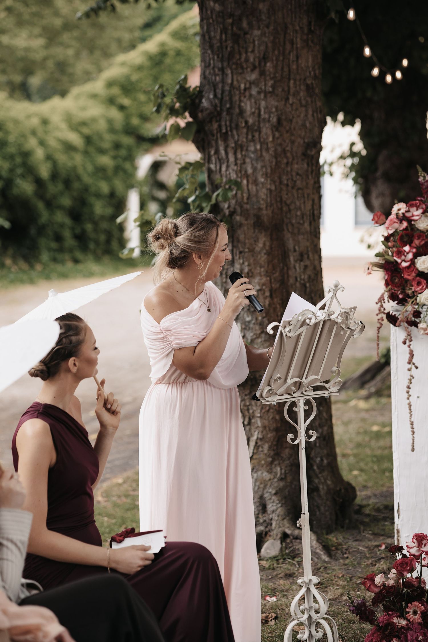 Bridesmaid in pink off-shoulder gown reading speech at outdoor wedding ceremony near floral arrangement.
