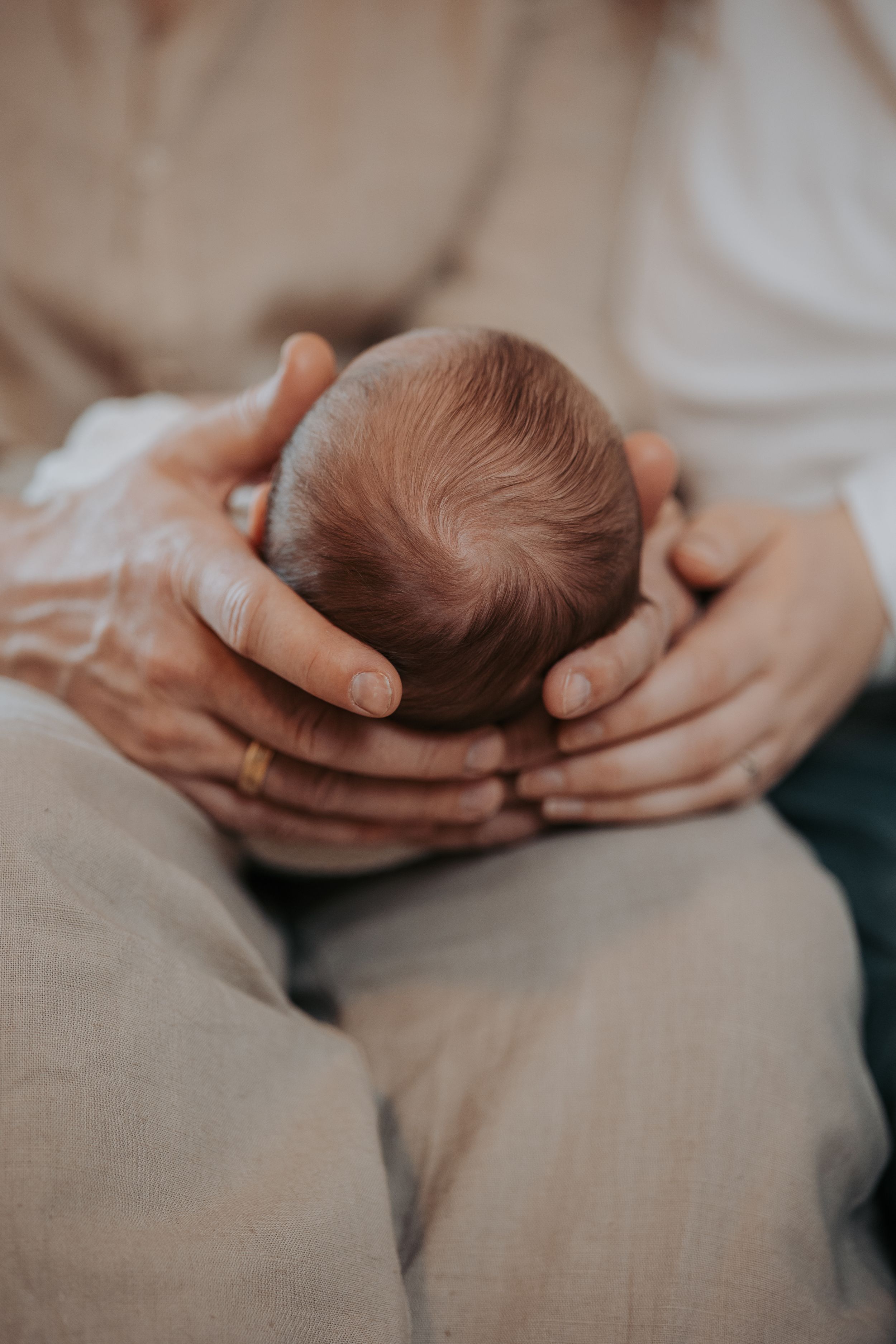 Gentle hands cradle a newborn baby's head showing soft brown hair in an intimate, muted-tone photograph.