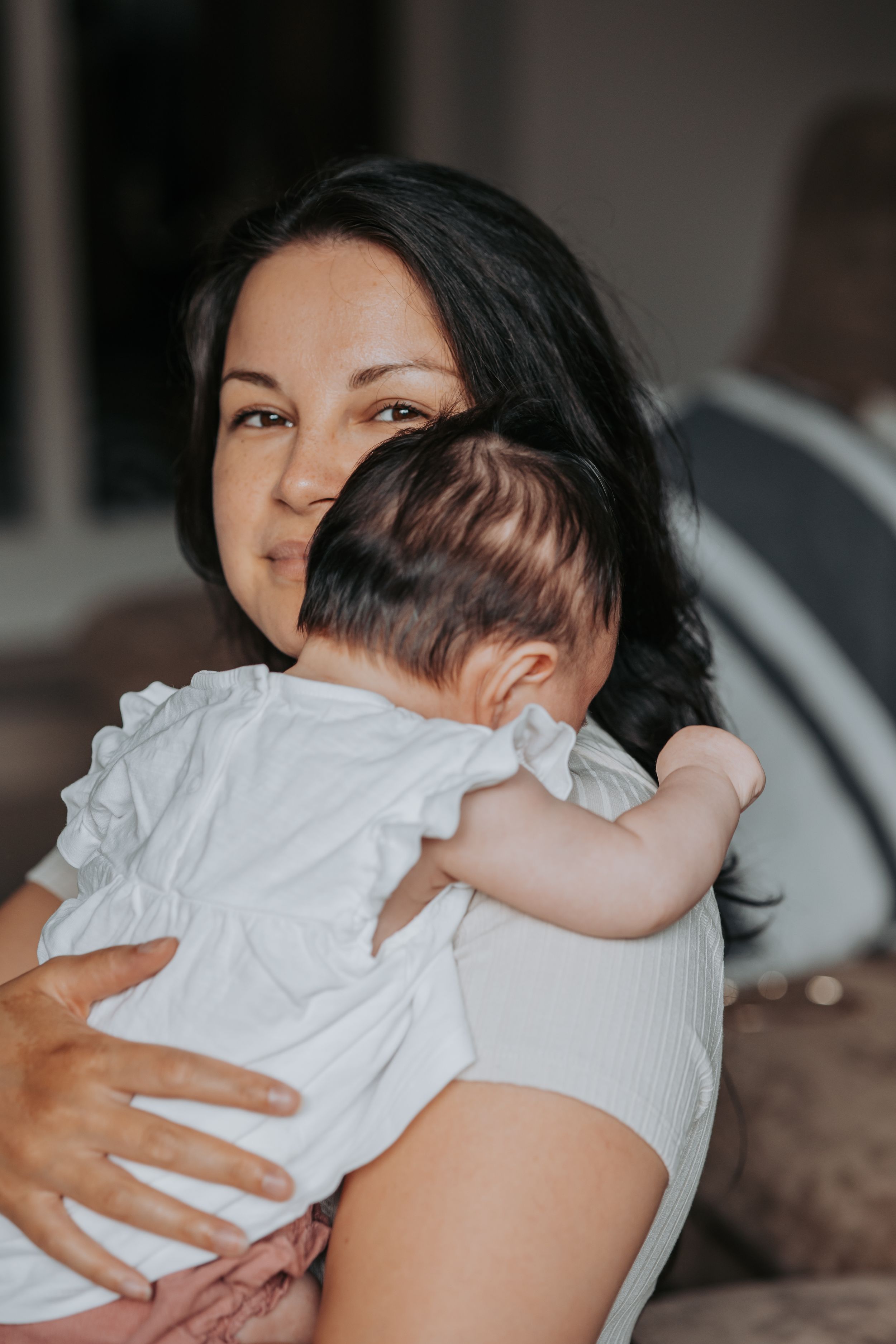 Parent in white shirt holds baby wearing white dress in tender indoor portrait.