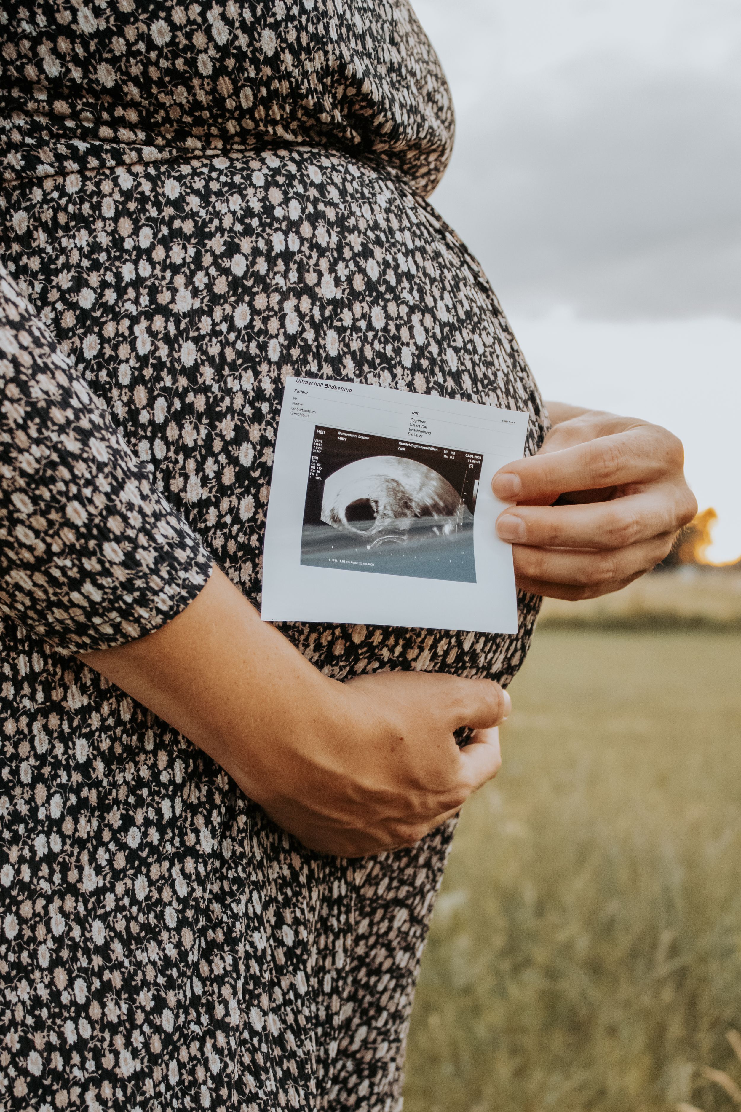 Pregnant woman in floral dress holds ultrasound photo against her belly while standing in grassy field on cloudy day.