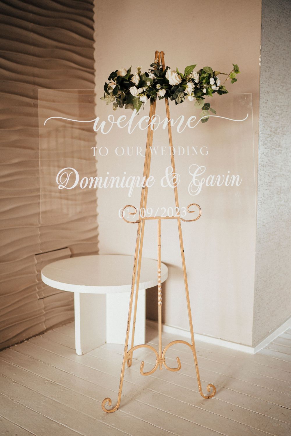 Elegant wedding welcome sign on a gold easel features calligraphy text and greenery decor against a neutral wall.