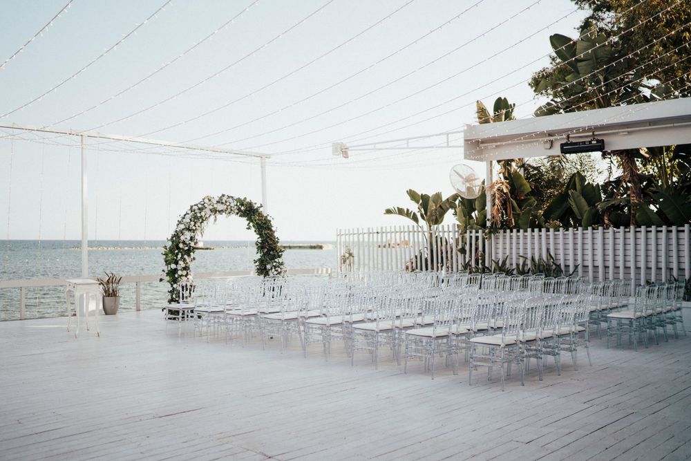 A beachfront wedding ceremony setup with a floral arch and clear ghost chairs overlooking the ocean at sunset.