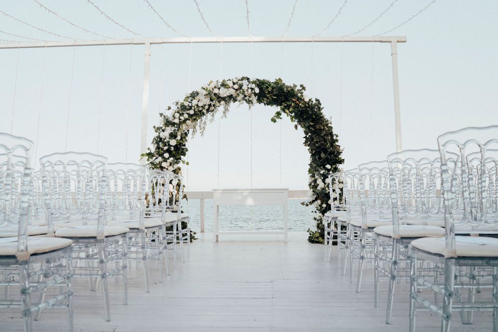 Floral wedding arch decorated with white flowers stands at the end of an aisle lined with clear ghost chairs.