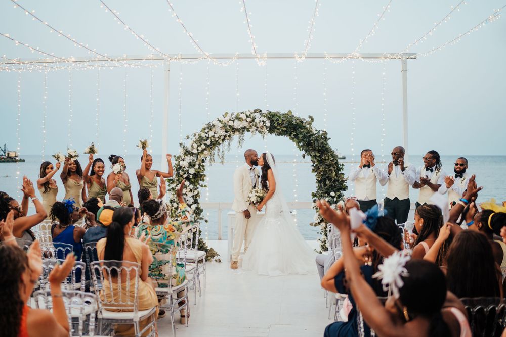 A destination beach wedding ceremony under a floral arch with guests capturing the special moment on a bright sunny day.
