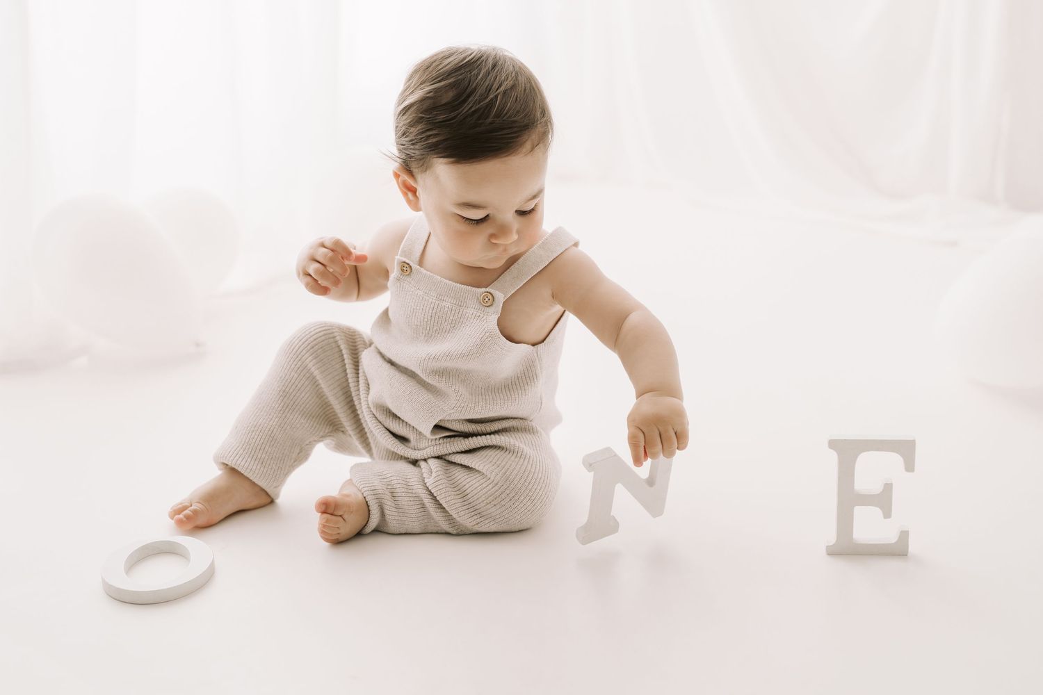 Bambino di un anno seduto in un set fotografico bianco, indossa un salopette beige e gioca con lettere di legno che compongono "ONE"