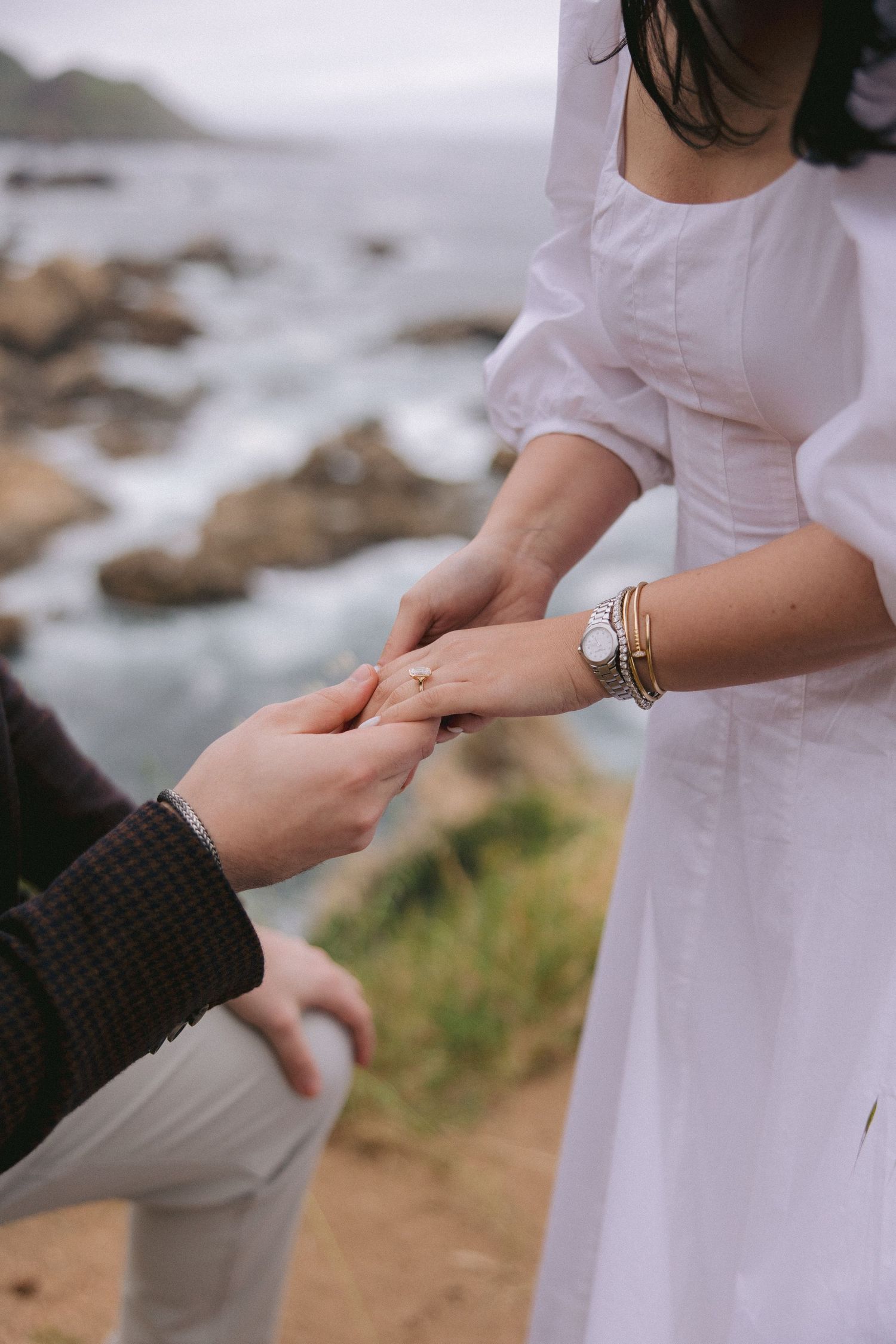 A couple holds hands on a rocky coastal cliff, with the bride wearing a flowing white dress and bracelet.