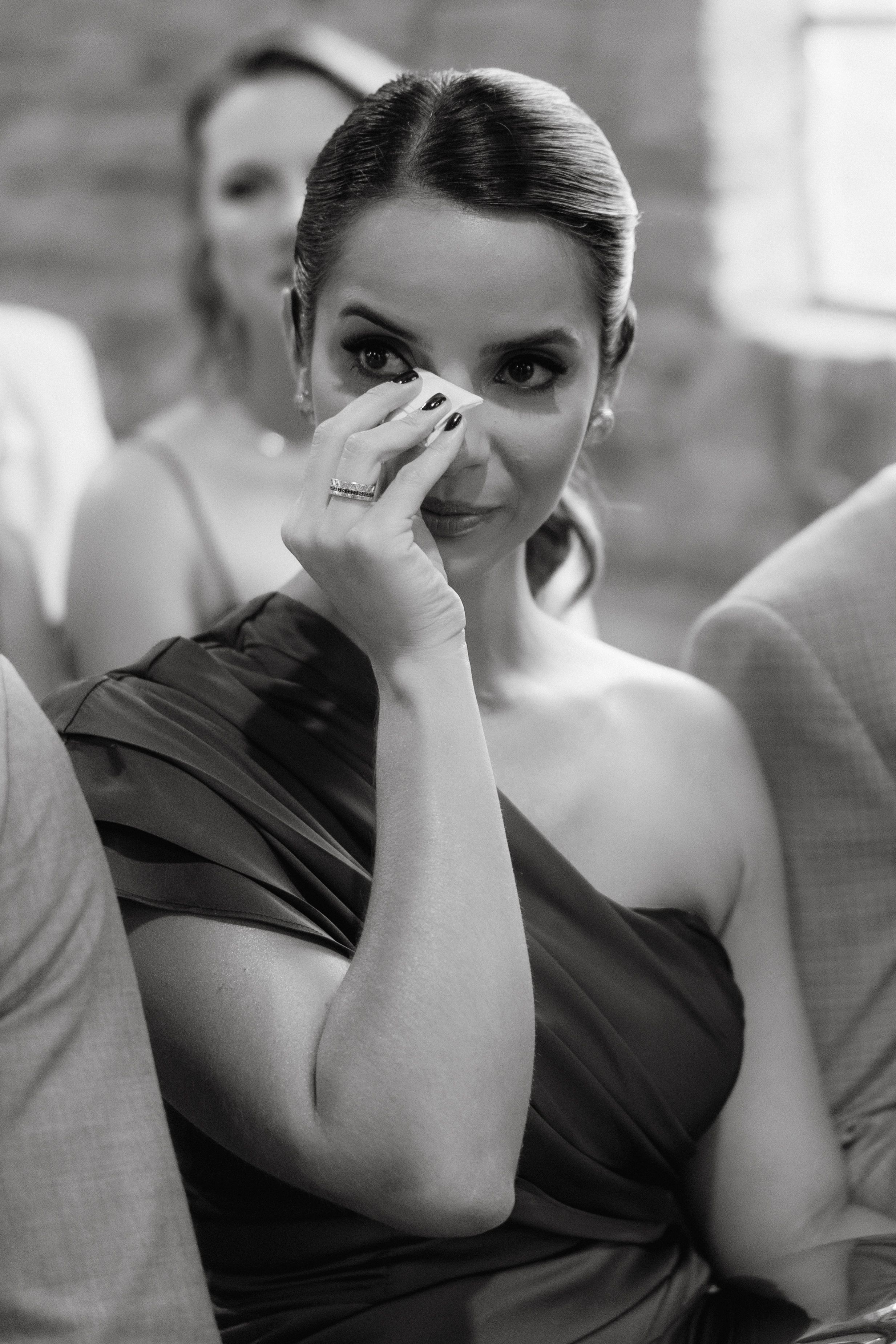 Emotional guest in one-shoulder dress wipes away tears during wedding ceremony.