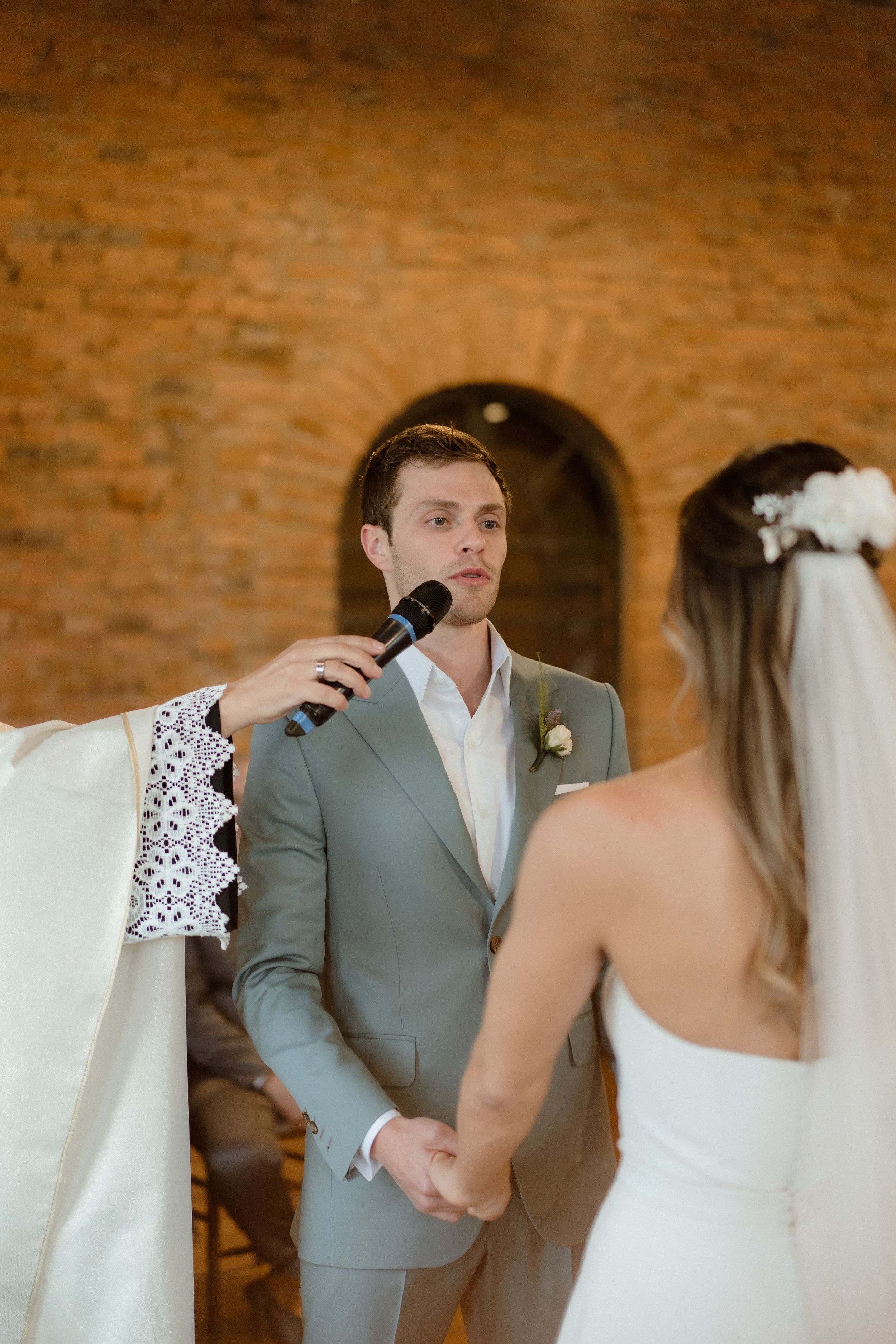 Groom speaks into microphone during wedding ceremony in rustic brick venue.