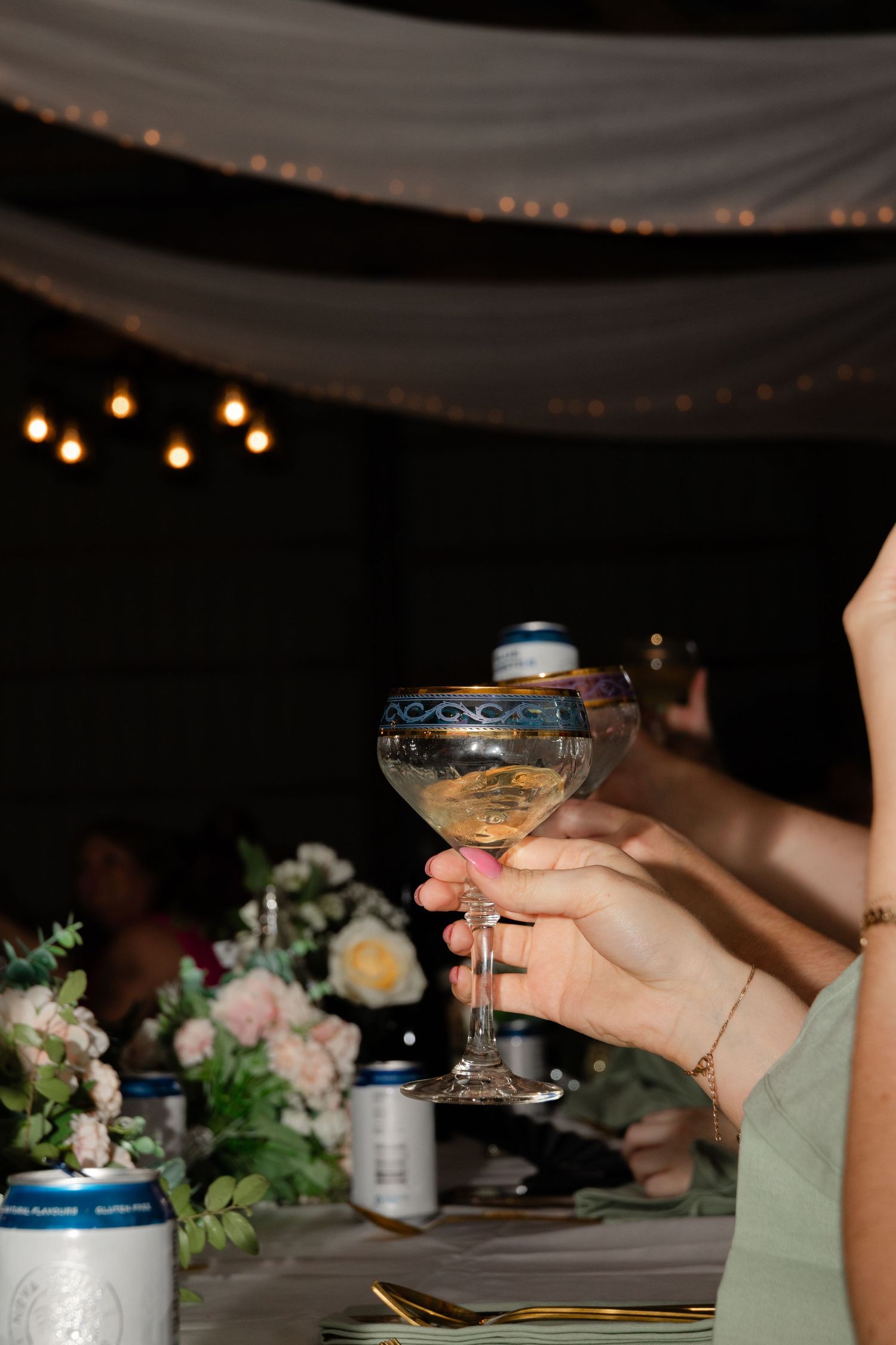Hand holding an elegant wine glass with floral arrangements and string lights visible in the background at an evening event.