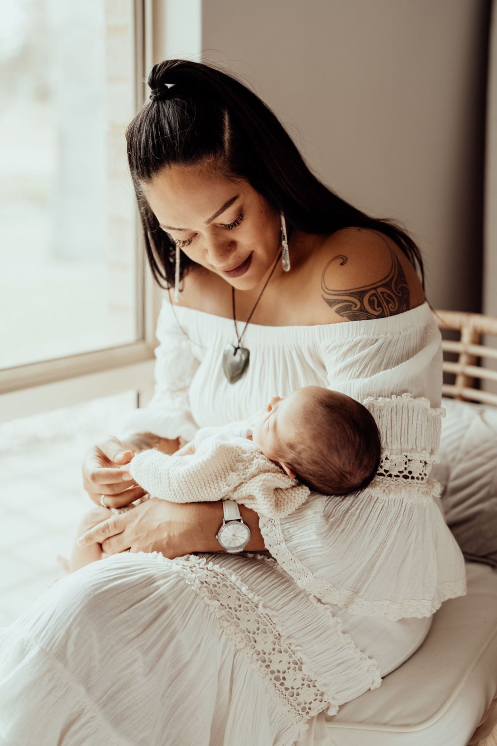 Tender moment between parent and newborn baby in white lace dress by window light.