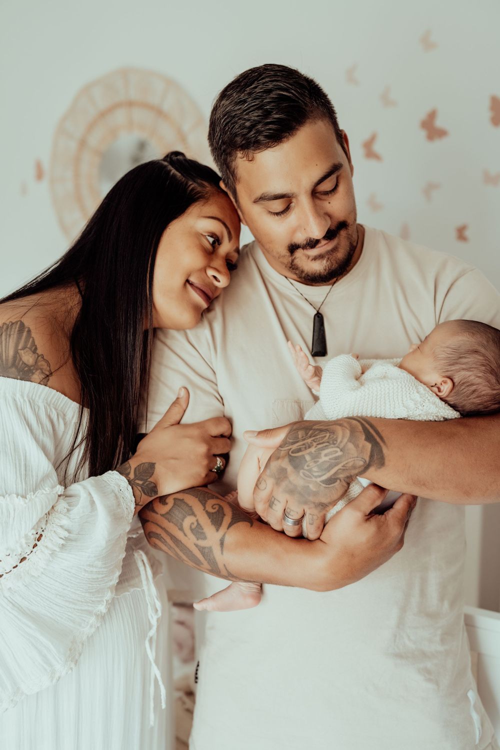 Family portrait with parents in white outfits cradling newborn baby with loving expressions.